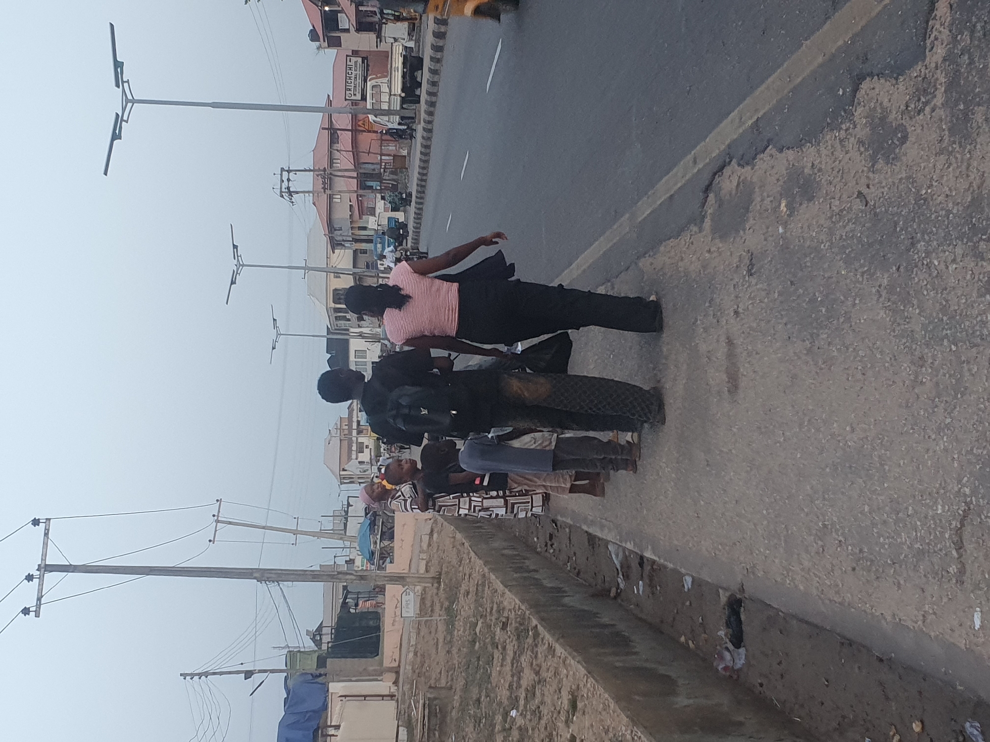 The image depicts a group of people standing on the roadside in Lokoja, Nigeria. They are near a major road with multiple lanes of traffic, and several buildings are visible in the background. A row of utility poles and streetlights line the road. Debris is scattered along the edge of the road.