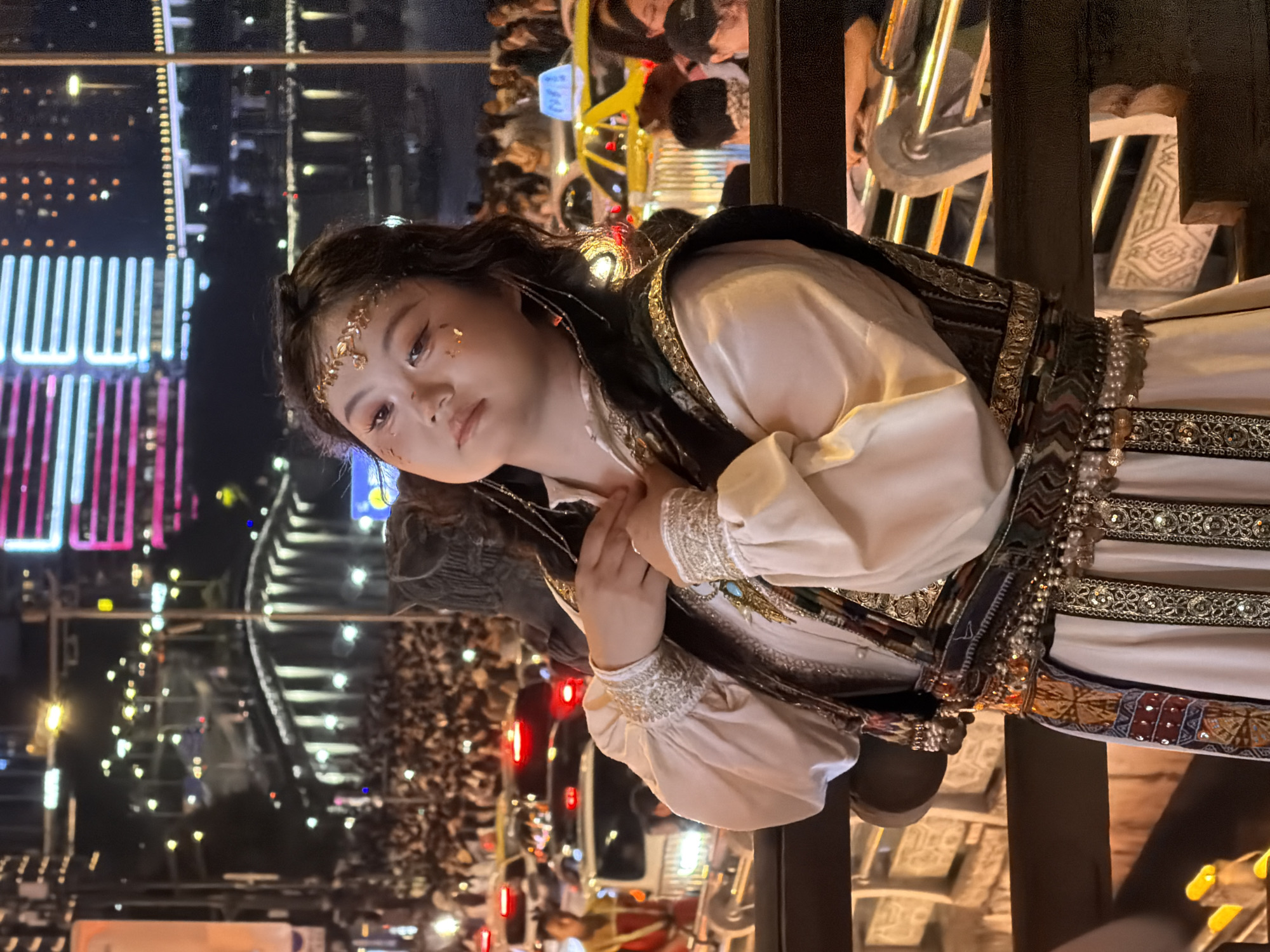 A young East Asian female, adorned in traditional costume with gold accents on her hair and face, is pictured at night in a crowded urban environment. She is positioned in the foreground, looking towards the camera with a slight tilt of her head. Her costume features a cream-colored blouse with ornate cuffs and a dark vest with gold embroidery. The background reveals a bustling street scene, possibly during a festival or event. Numerous people are visible, suggesting a large gathering. In the distance, illuminated buildings and traffic lights create a vibrant, colorful backdrop characteristic of a city at night. The scene is likely located in Yuzhong District, China, given the architectural style and urban setting.