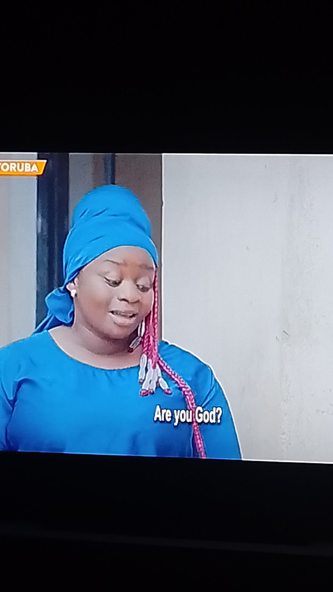 The image shows a medium shot of a Black woman wearing a bright blue headscarf and a matching blue dress. Her hair is braided with pink extensions adorned with white beads. She is looking down and to her right, with her mouth slightly open as if speaking. Text overlays the bottom right of the image, reading "Are you God?". A logo for "YORUBA" is visible in the top left corner. The background is a plain, off-white wall. There are no discernible elements in the image to suggest a specific location within Lokoja, Nigeria.