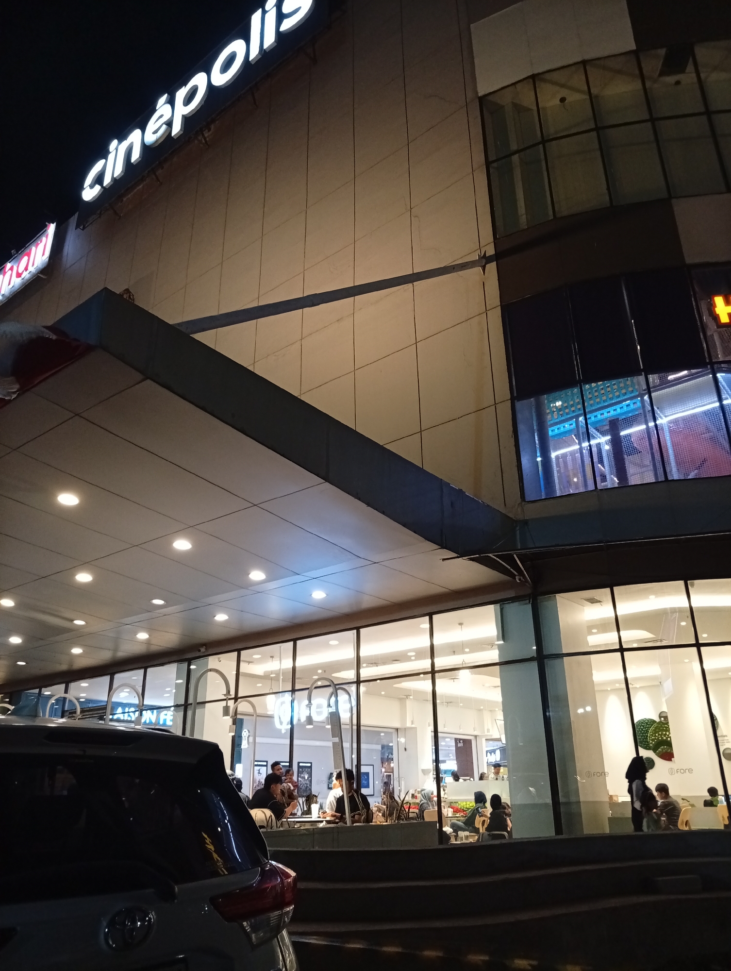 The image depicts the exterior of a Cinepolis cinema at night, likely located in Sidoarjo, Indonesia, as suggested by the text on the building. The prominent signage "cinépolis" is illuminated in white. The building features a modern architectural design with large glass windows and a beige tiled facade. Inside the cinema, visible through the windows, are people seated at tables, possibly in a lobby or food court area. Some individuals are standing, and one appears to be holding a baby. The scene is well-lit from within the building and by overhead lights, creating a warm glow. In the foreground, the rear of a dark-colored SUV is visible, partially obscuring the entrance. The overall impression is that of a busy commercial area at night, with a focus on entertainment and dining.