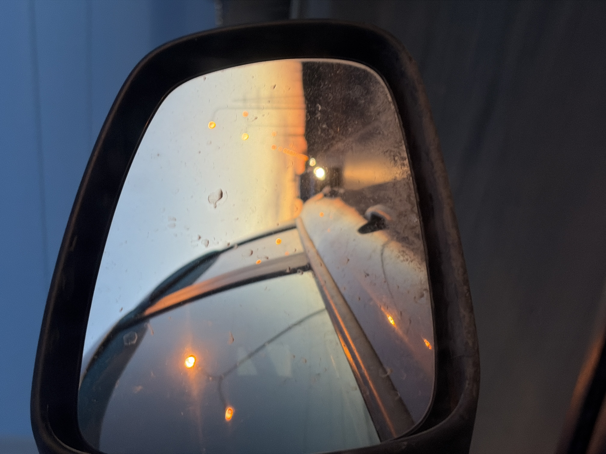 The image shows a car's side mirror reflecting a scene at dusk or dawn. The reflection includes the side of the vehicle the mirror is attached to, with faint orange lights suggesting streetlights or other vehicles. There's also a blurred reflection of what appears to be a passing car with headlights. The mirror surface has water droplets. The surrounding environment visible outside the mirror is dark, indicating low-light conditions. The location in the city of Chaikovsky, Russia, is not determinable from the image.