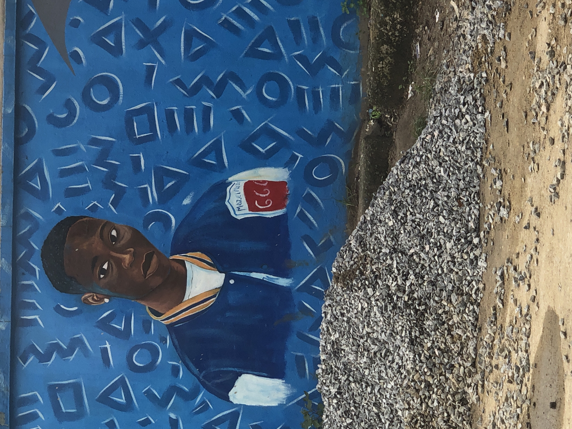 The image shows a mural of a young Black male wearing a blue bomber jacket with white and orange trim, and a white t-shirt. He has short, dark hair and is wearing an earring. The background of the mural is a vibrant blue with white geometric shapes, including triangles, circles, and zig-zag lines. On the left breast of the jacket, there is a red emblem with the text "Mozelife" and the number "666." The mural is painted on a concrete wall. In front of the wall, there is a large pile of grey gravel. The ground in front of the gravel is dirt with scattered stones. The scene appears to be outdoors in an urban environment, possibly in Ikeja, Nigeria.