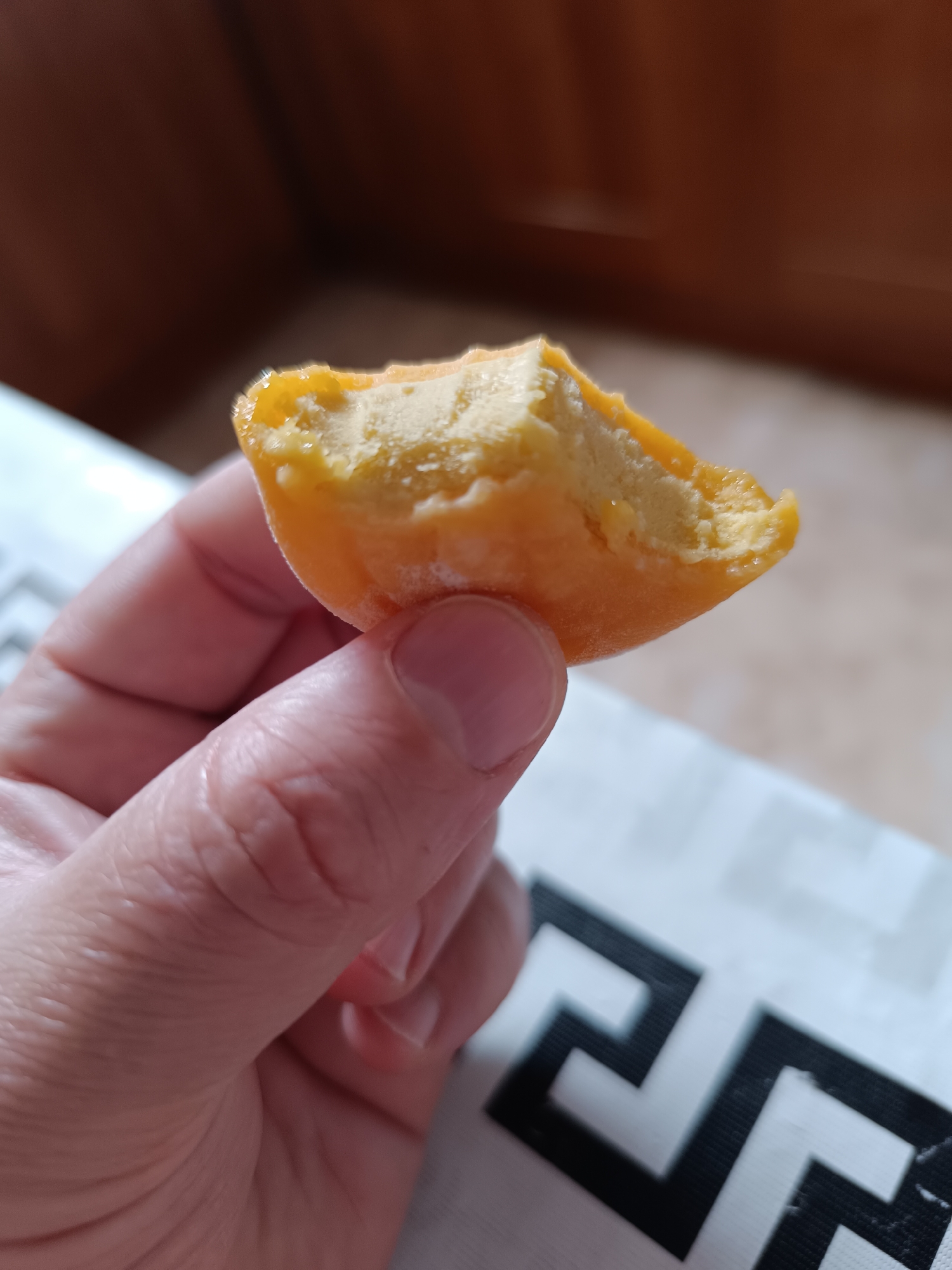 A close-up shot shows a person's hand holding a piece of bright orange mochi ice cream with a bite taken out of it. The mochi has a slightly textured, powdery exterior and a creamy, pale yellow ice cream filling that appears to be melting. The background is blurred, with a dark brown surface above and a white surface with a black geometric pattern below. The scene appears to be indoors.