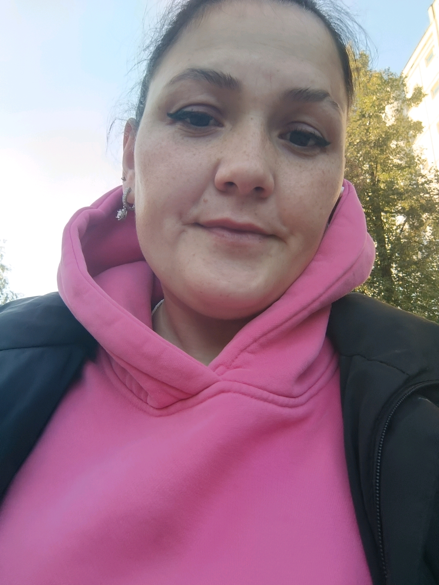 A medium-shot, outdoor selfie shows a woman with her head slightly tilted, looking directly at the camera. She has dark hair pulled back, light skin with freckles, and is wearing winged eyeliner. She has a small stud earring in her left ear. She is wearing a bright pink hooded sweatshirt, and a dark jacket over it. Behind her, the sky is a pale blue, with a few scattered clouds. To the right, the top of a building and lush green trees are visible. The image is likely taken in Chaikovsky, Russia, given the context of other images from the same source.
