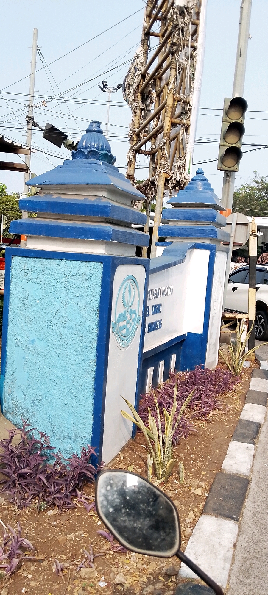 The image depicts a decorative gateway structure with blue and white accents, likely marking an entrance in Depok, Indonesia. The gateway features tiered, pyramidal roofs topped with finials, and a white wall with blue trim. A circular emblem or seal is visible on the left pillar, and text, partially obscured, appears on the white wall, possibly indicating a location name. In the foreground, a motorcycle's rearview mirror is reflected, showing a dirty, slightly smudged surface. Surrounding the gateway are various plants, including low-lying purple foliage and taller, spiky green plants. The ground is covered with soil and small stones. In the background, electrical wires crisscross overhead, and a traffic light with three visible signals is mounted on a pole to the right of the gateway. A portion of a white vehicle is visible behind the gateway structure. The overall scene suggests an outdoor urban or suburban environment.