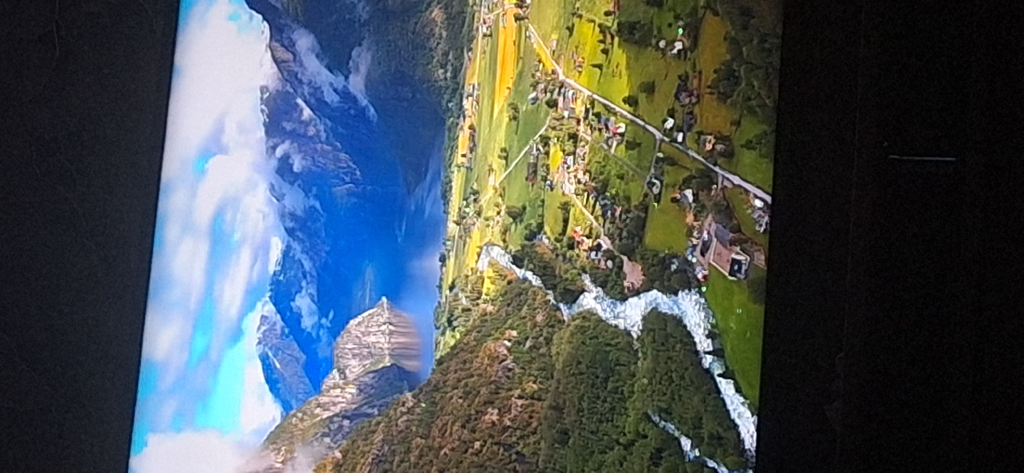 This is a high-angle, aerial view of a scenic landscape. A vast, deep blue body of water, likely a fjord or large lake, dominates the mid-ground, flanked by steep, rocky mountains on the left and softer, more verdant mountains on the right. White clouds drift across the sky and cling to the mountain peaks. In the foreground, a winding river with white, turbulent water flows from the left towards the center. Adjacent to the river, on the right side, is a rural settlement with numerous houses and buildings scattered across green fields and interspersed with trees. The landscape exhibits lush green vegetation on the mountainsides and in the valley, with patches of yellow and orange suggesting cultivated fields. The overall impression is one of natural beauty and tranquility, with a small human presence integrated into the environment. This scene does not appear to be located in Bila Tserkva, Ukraine; it depicts a mountainous, fjord-like region.