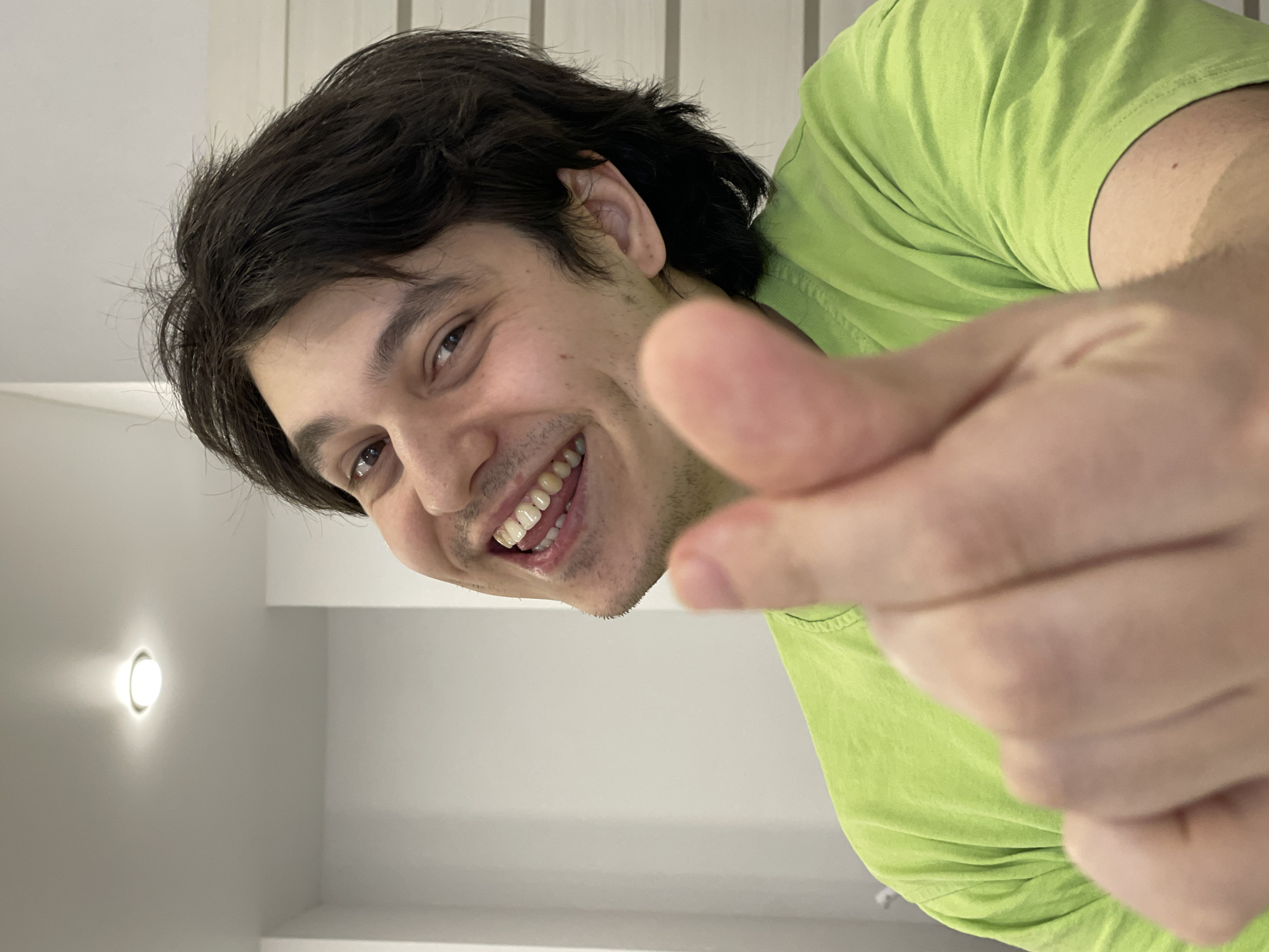 A man is positioned closely to the camera. He is smiling and pointing directly at the viewer. He has dark hair and is wearing a lime green t-shirt. The background is a white-walled room with a round ceiling light. There is no indication that the photo was taken in Izhevsk, Russia.