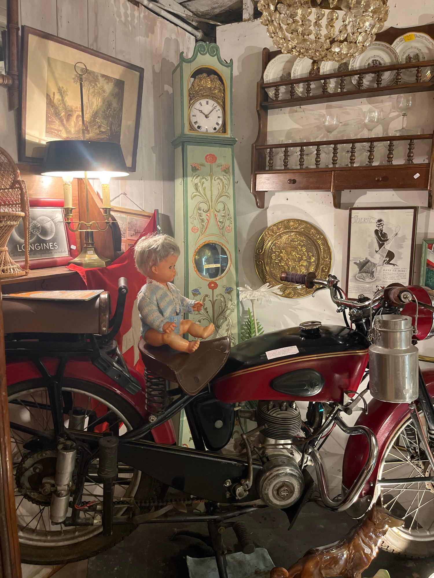 In a cluttered shop in Yvoire, France, a vintage red motorcycle is parked indoors. A doll, dressed in a blue and white striped outfit, is seated on the motorcycle's saddle. Behind the motorcycle, a tall, slender grandfather clock with ornate green and red floral designs stands against a white-paneled wall. To the left of the clock, a framed landscape painting is displayed above a table with a lamp, a brass candelabra, and a framed advertisement for Longines watches. Further to the right, a wooden shelf unit holds decorative plates and glassware. A framed vintage poster featuring a woman and a rooster is also visible.