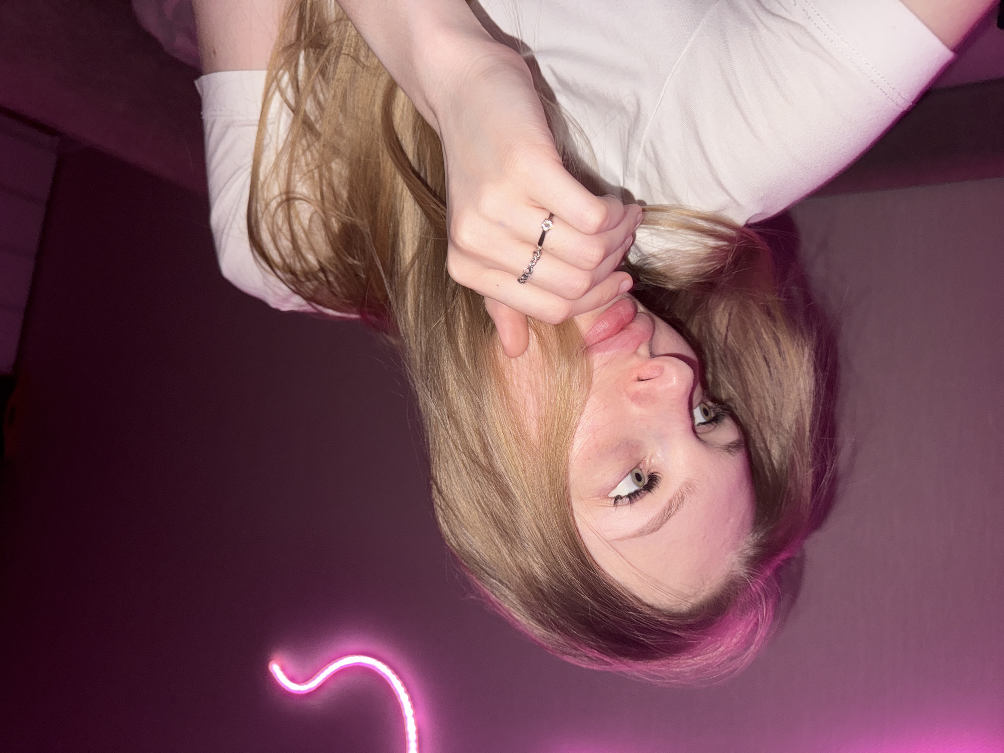 The image shows a close-up of a young woman with blonde hair, looking to her left. She is wearing a white t-shirt and has a ring with small stones on her left hand, which is near her mouth. A pink neon light is visible in the background. The scene is indoors, with a pink-hued wall and a dark background. There is no information to suggest the location is in the city of Perm, Russia.