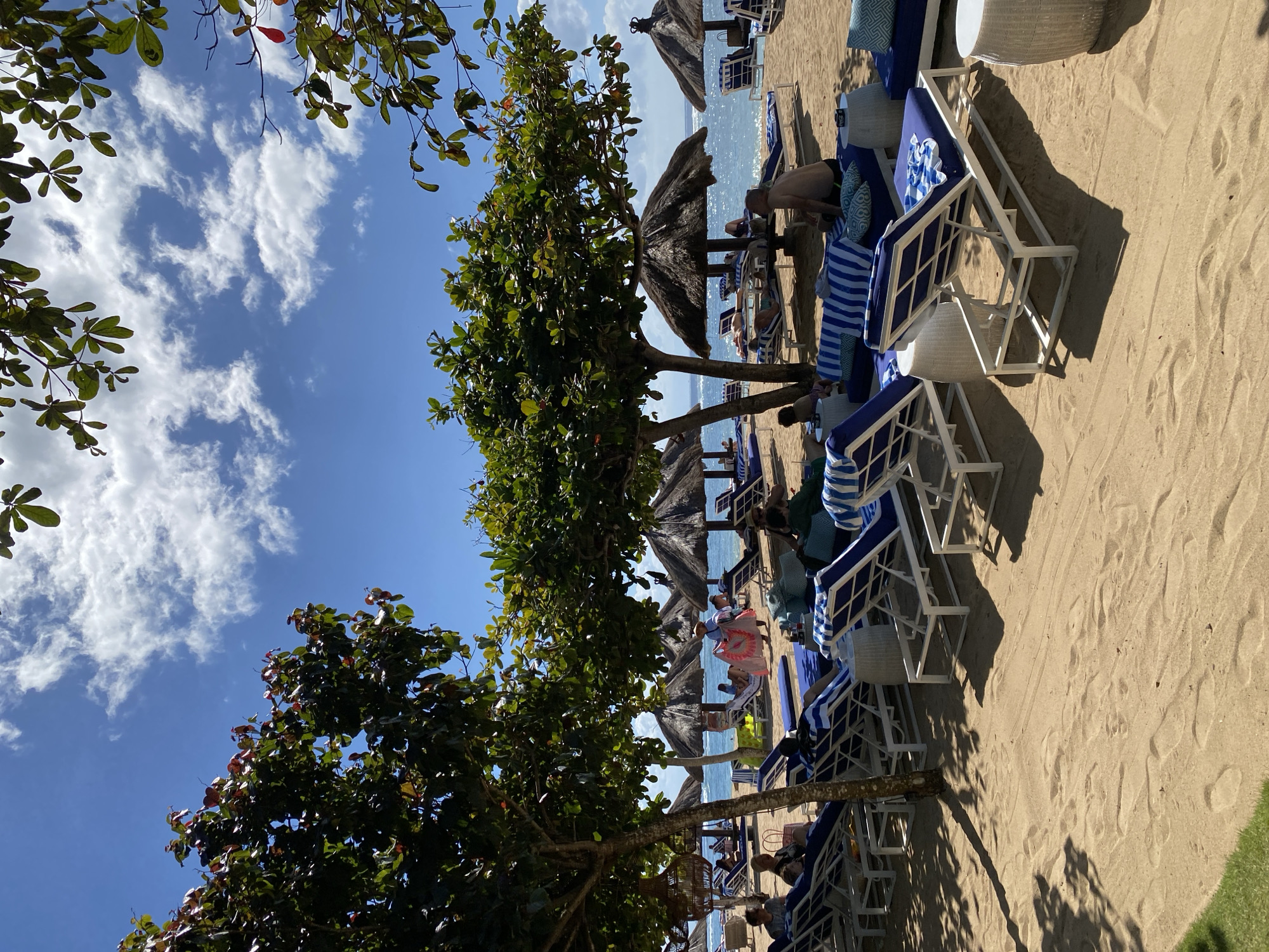 The image depicts a beach scene in Kuta Selatan, Indonesia. Several rows of white-framed beach loungers with blue cushions are arranged on the sand. Some loungers are occupied by people, some of whom are partially visible. In the background are thatched umbrellas providing shade. The turquoise ocean is visible between the umbrellas and the beach. Trees with green foliage frame the scene on both sides. Above, the sky is blue with scattered white clouds.