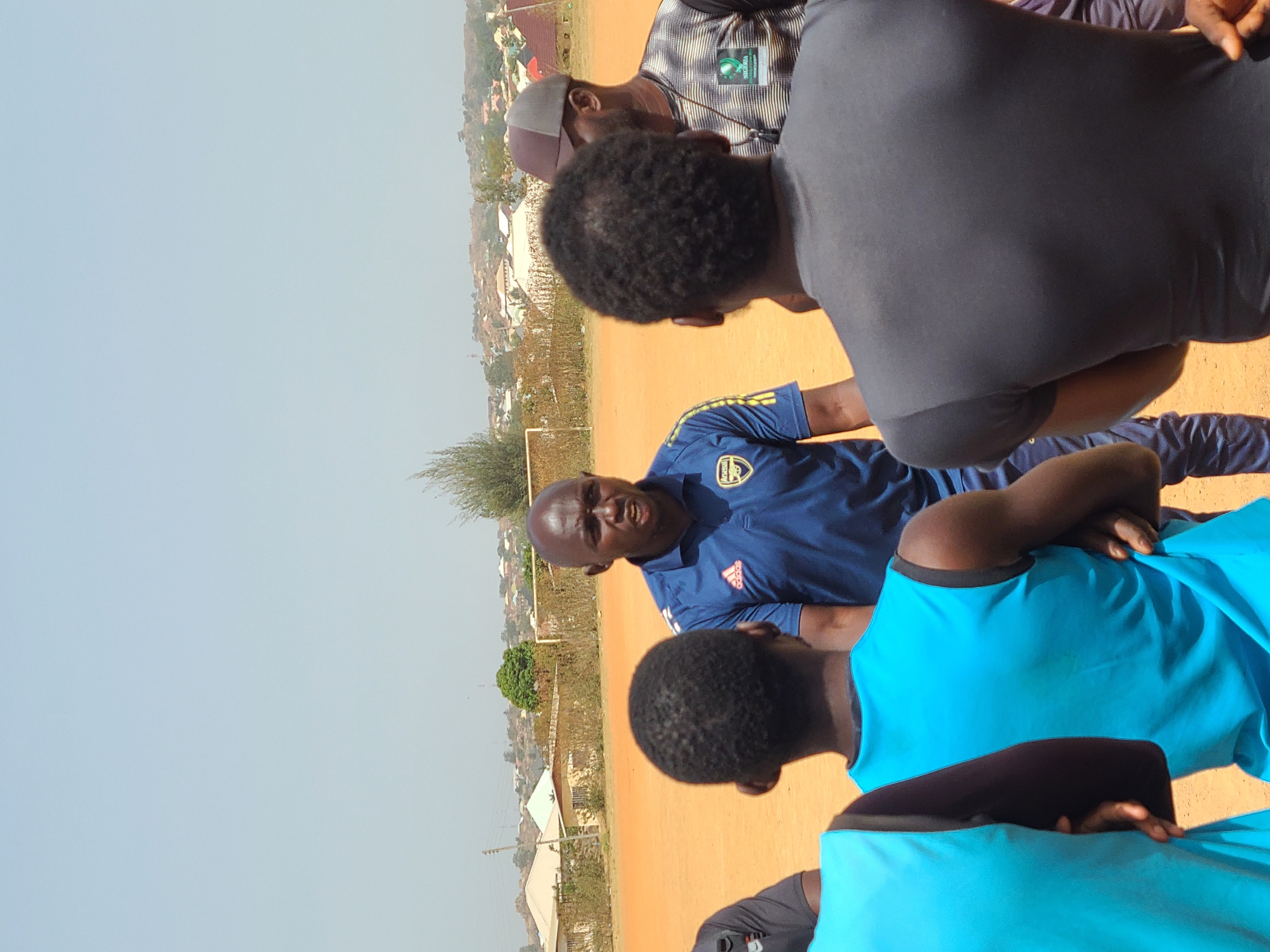 This outdoor image, captured in Barikin Jirga Summa, Nigeria, depicts a group of men gathered on a reddish-brown, unpaved ground, likely a sports field or open area. The sky above is clear and light blue, indicating bright daylight.
In the center, a man with a bald head is looking towards the viewer, seemingly addressing the group. He is wearing a blue short-sleeved athletic shirt featuring the "Adidas" logo and word, along with what appears to be an "Arsenal" emblem on the chest. His expression suggests he is actively speaking or instructing.
Surrounding him are other men, some viewed from behind or partially. In the immediate foreground, a person with dark, curly hair is seen from the back, wearing a black t-shirt. Another individual, also seen from behind, wears a light blue t-shirt. Further back and to the right, a man in a striped shirt and a light-colored cap is visible, with a green badge pinned to his shirt, which has an unreadable logo and text.
The setting is an open, dusty field, with sparse vegetation. In the background, numerous buildings with light-colored roofs are visible, along with some trees, suggesting a village or town bordering the training area. The scene conveys a sense of a focused discussion or coaching session taking place under clear, sunny weather conditions.