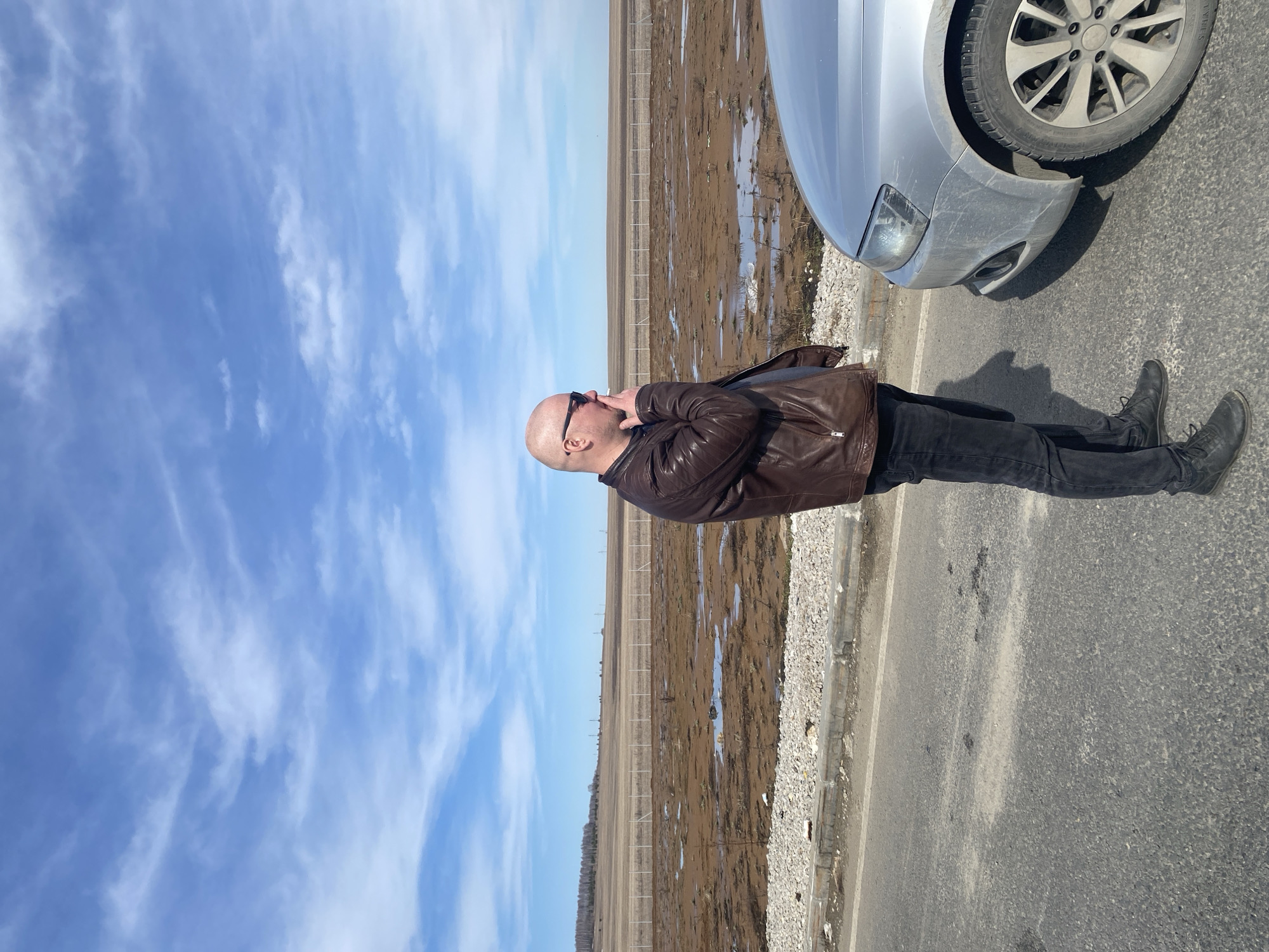 A bald, light-skinned man wearing a brown leather jacket, black pants, and black boots stands on the asphalt shoulder of a road in Kliuchiki, Russia. He has sunglasses on and his left hand is near his face. The front of a silver car is partially visible to his right. Beyond the shoulder of the road, there's a muddy, barren field enclosed by a fence. The sky is blue with scattered white clouds.