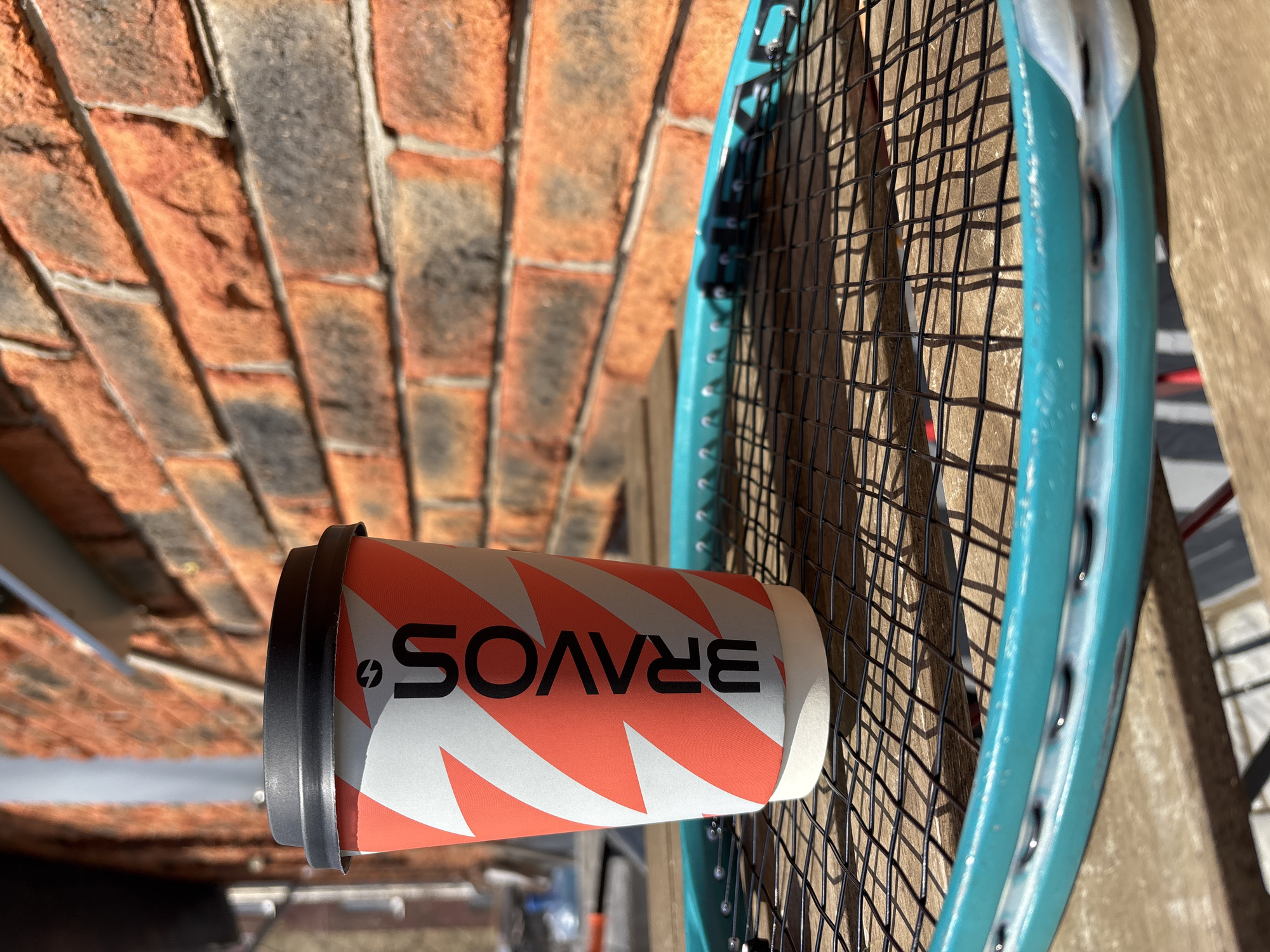 An angled, close-up photograph captures a paper cup and a tennis racket positioned outdoors in Chaikovsky, Russia, under bright daylight. The scene is well-lit by direct sunlight, creating strong shadows and indicating a clear, sunny day.
The central subject is a white paper cup adorned with a dynamic coral-orange zig-zag pattern. A black plastic lid covers one end of the horizontally oriented cup. Printed prominently on the cup in black capital letters is the word "BRAVOS," accompanied by a stylized lightning bolt or arrow symbol to the left of the 'S'. In the image's orientation, the text appears upside down.
Partially visible on the right is a tennis racket, featuring a vibrant teal or light blue frame. Its black strings form a tight grid. The racket appears to be resting against a light brown, possibly wooden, surface, which is also visible on the far right.
The background consists of a textured brick wall, showcasing a mix of red, orange, and gray bricks arranged in a pattern, possibly an archway. No people are present, and no specific activity is taking place, presenting a still life composition of these items in an outdoor setting.