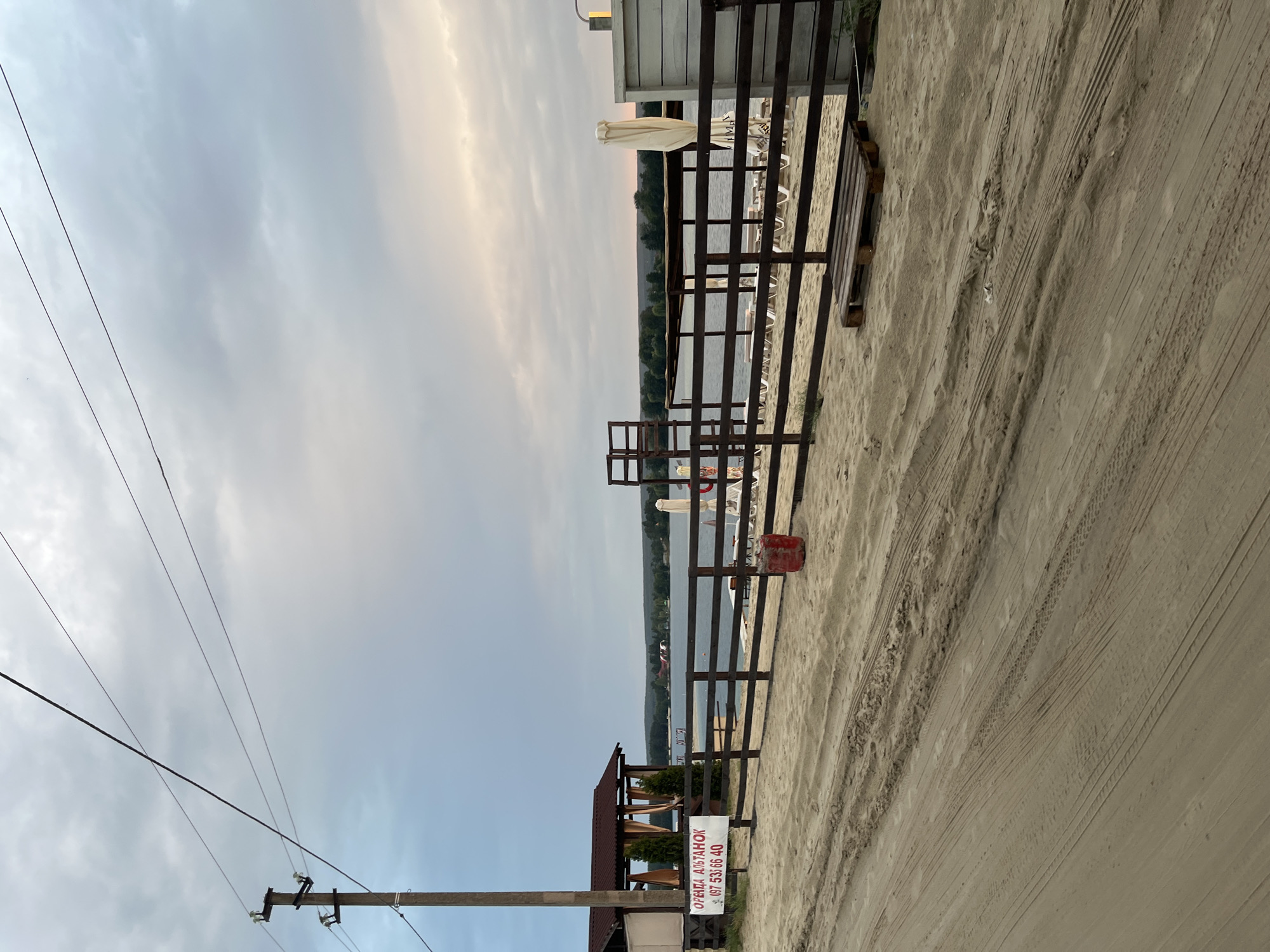 The image depicts a beach scene by a lake, likely on the outskirts of Bezliudivka, Ukraine, under a cloudy sky. In the foreground, a sandy path with tire tracks runs diagonally across the frame. To the left, a wooden utility pole with wires stands prominently. Adjacent to it, a covered pavilion with brown curtains and green bushes is visible. A white sign attached to a wooden fence reads "ОРЕНДА АЛЬТАНOK" and a phone number "097 53 66 40". The fence encloses a beach area with numerous lounge chairs and umbrellas, some of which are folded. In the middle ground, the lake stretches towards a tree-lined horizon. The water is calm, reflecting the overcast sky. A red cylindrical object, possibly a bin or buoy, is located near the fence. To the right, a section of a white wooden structure is partially visible. The overall impression is one of a quiet, possibly closed, beach resort.