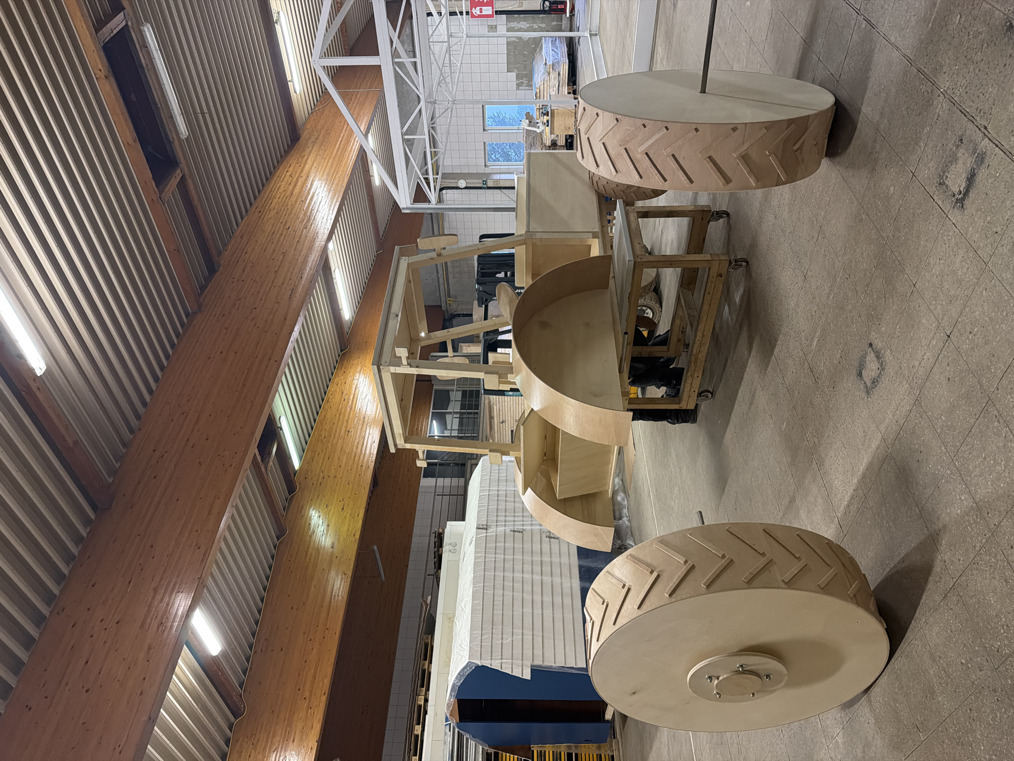An unfinished wooden tractor structure is in the foreground, situated on a concrete floor within a workshop. The tractor has large, treaded wooden wheels and a frame that resembles a cab. To the left, a blue and white object covered in plastic sheeting is partially visible, possibly another construction element or vehicle. In the background, a white metal frame structure, resembling a greenhouse or shed, is present. The workshop ceiling features exposed wooden beams and fluorescent lighting. The scene is located in a workshop in Leudelange, Luxembourg.