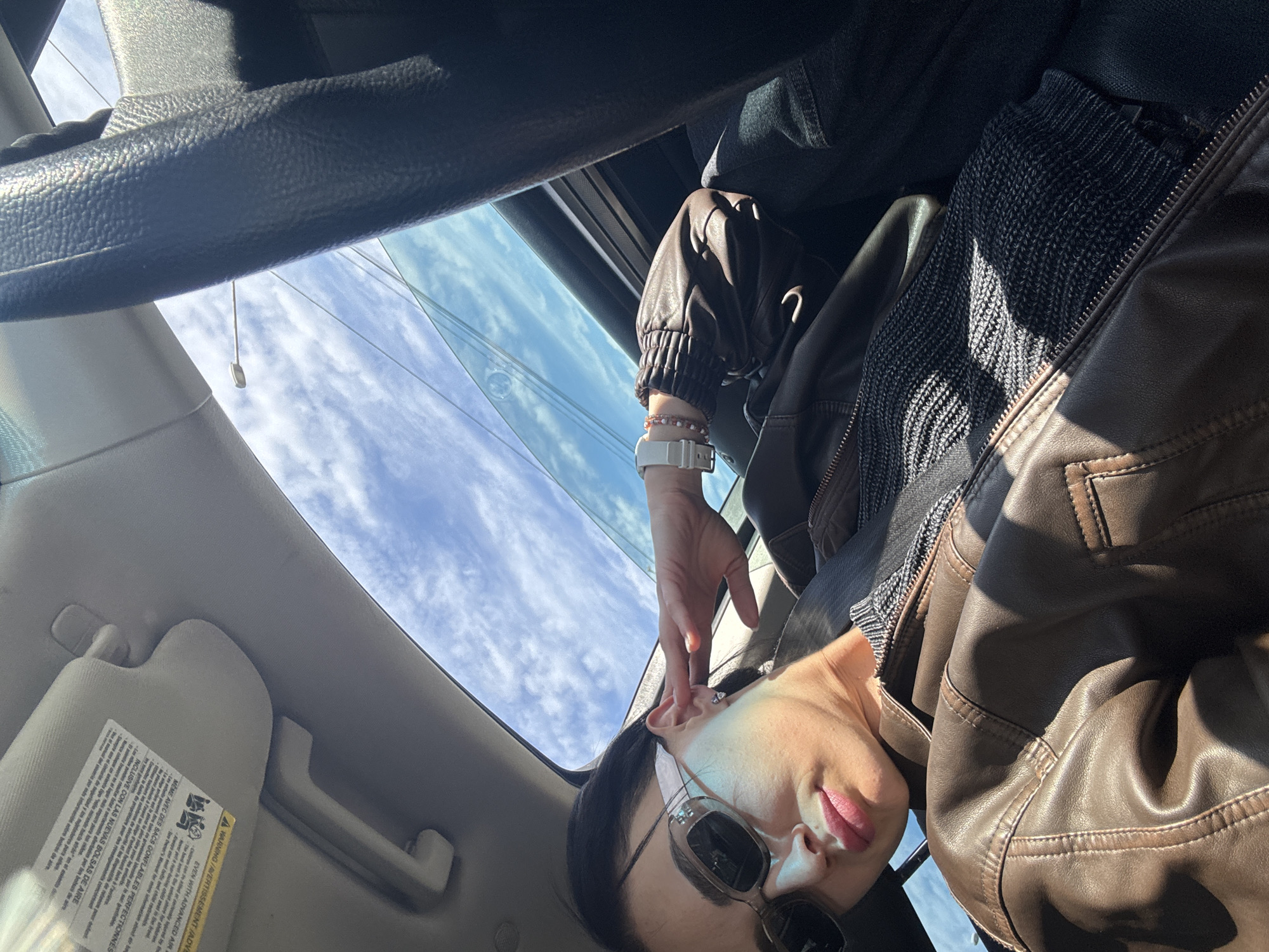 A person is sitting inside a car, seemingly under a clear, partly cloudy sky visible through a sunroof. The person is wearing sunglasses and a brown leather jacket with a textured, potentially knit, inner layer. The wrist of one arm is visible, adorned with a watch and a bracelet. A warning label is affixed to the sun visor of the car. It's impossible to determine if the car is in Tbilisi, Georgia.