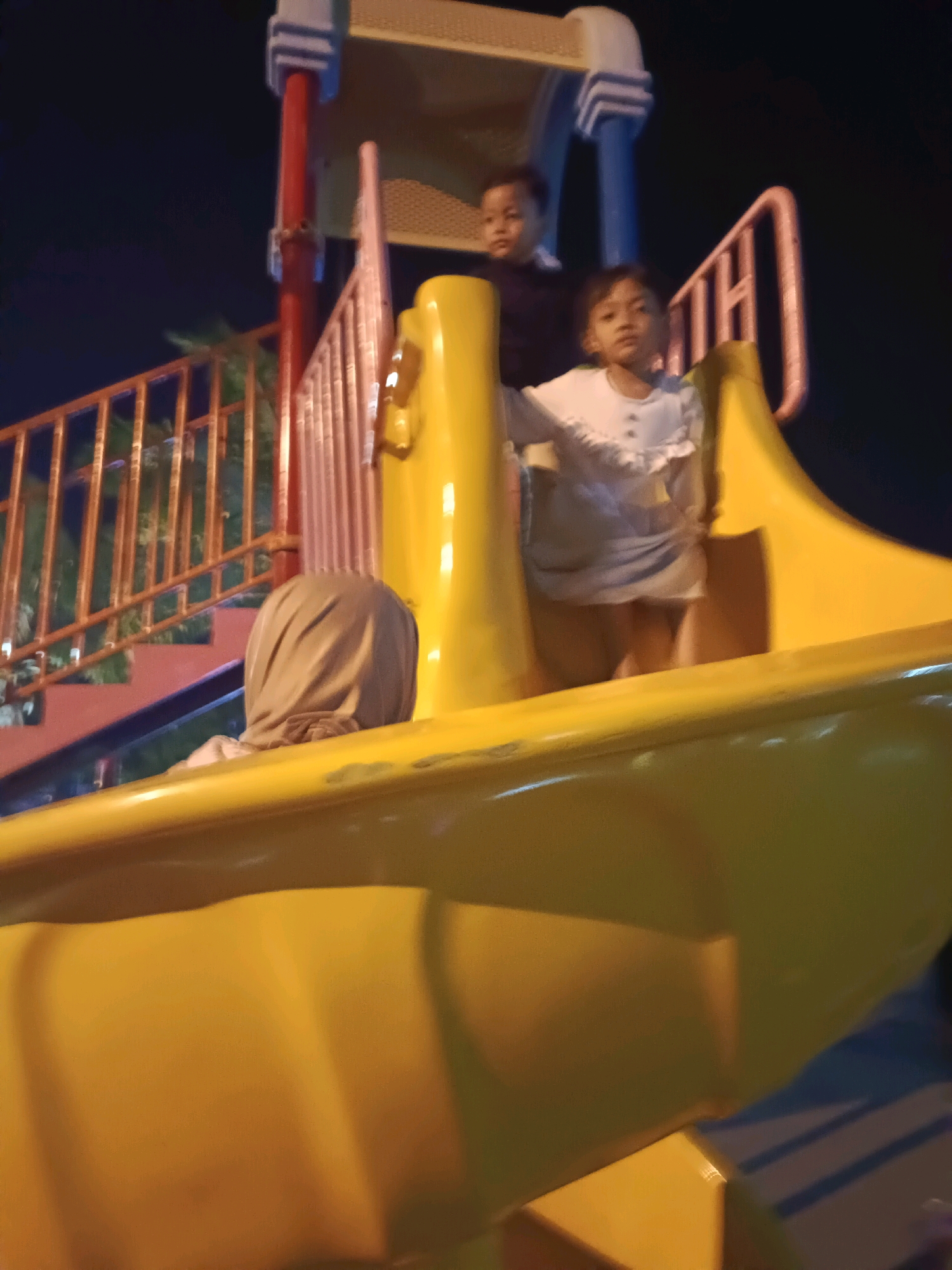 The image depicts a playground at night in the city of Lamongan, Indonesia. In the foreground, a large, yellow plastic slide curves into view. On the slide, a child wearing a light-colored top is positioned facing forward. Behind this child, another child, dressed in darker clothing, is also on the slide, looking to the left. Partially visible at the bottom left of the slide is a person wearing a headscarf. The playground structure features red and blue supports and a metal railing. The background is dark, indicating it is nighttime, with some out-of-focus greenery visible behind the railing.
