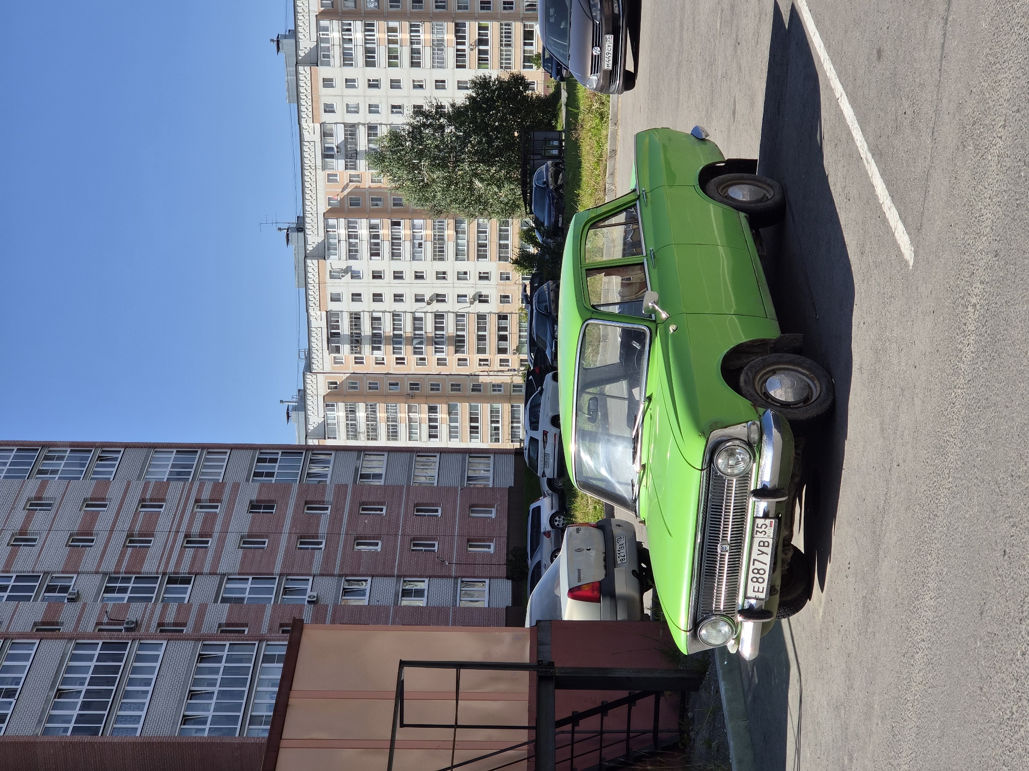 A bright green, vintage sedan is parked in a lot in front of a brick and light-colored apartment building in Cherepovets, Russia. The car, a Moskvitch 412 or similar model, has a chrome grille and bumpers, and its license plate reads "E887YB 35". The car is angled slightly towards the right side of the frame, with its front tires visible. In the background, other vehicles are parked, including a silver sedan and a dark SUV. The apartment buildings are tall and modern, with many windows. A large tree stands between the buildings and the parking lot. The sky is clear and blue, indicating a sunny day. The parking lot is paved with asphalt, and a white line marks a parking space. The overall scene depicts a typical urban environment in Russia.