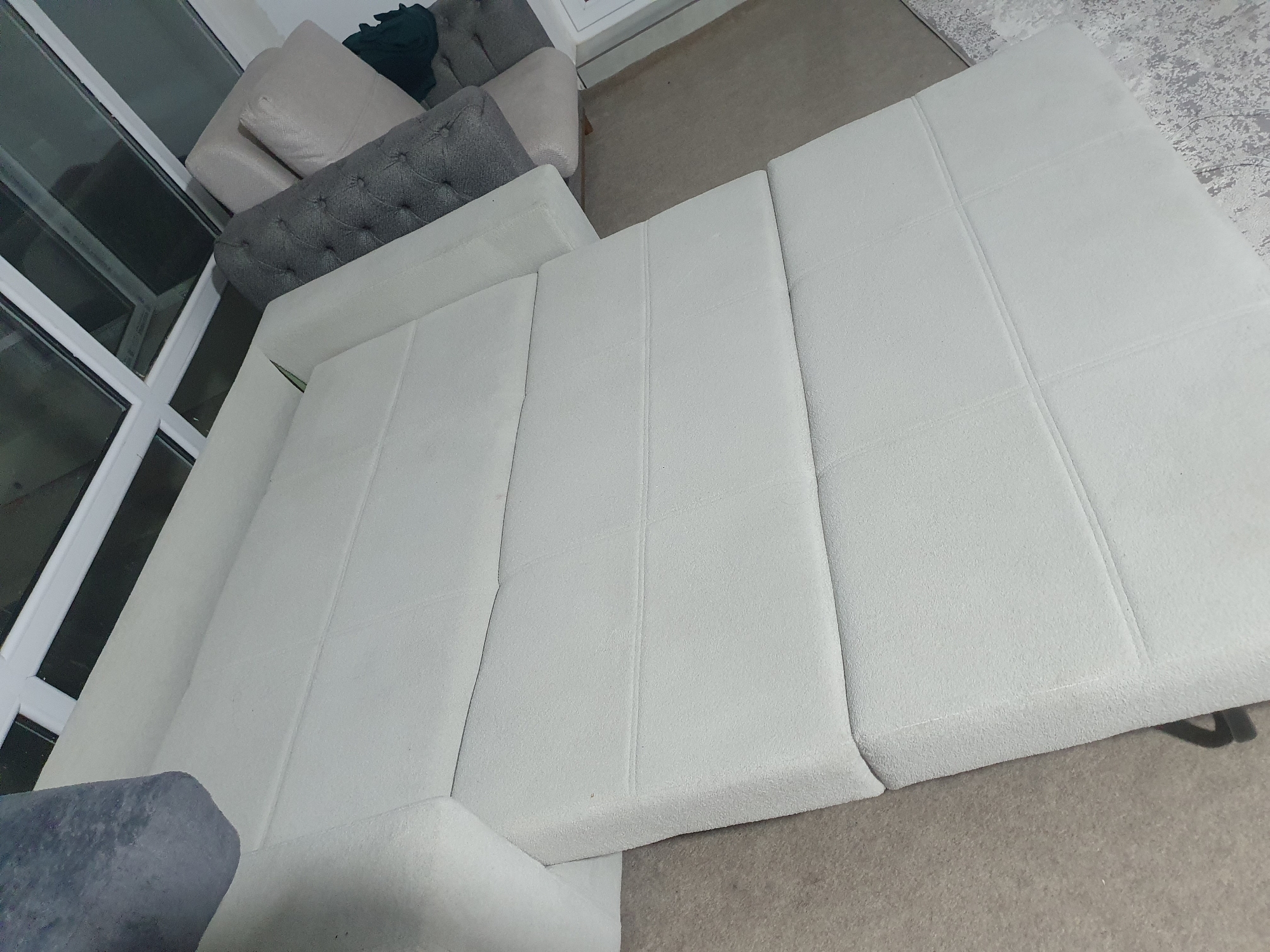 The image shows a sectional sofa in a room. The sofa is light-colored and has a textured fabric. It appears to be in good condition. There are some decorative pillows on the sofa, one of which is tufted and gray. The room has carpeted floors and a large window with a white frame. There is also another sofa or chair visible in the background, upholstered in a light color. The overall impression is one of a modern and comfortable living space. The image does not provide any specific information about the city of Kheyrabad, Iran, or any actions, interactions, or people.