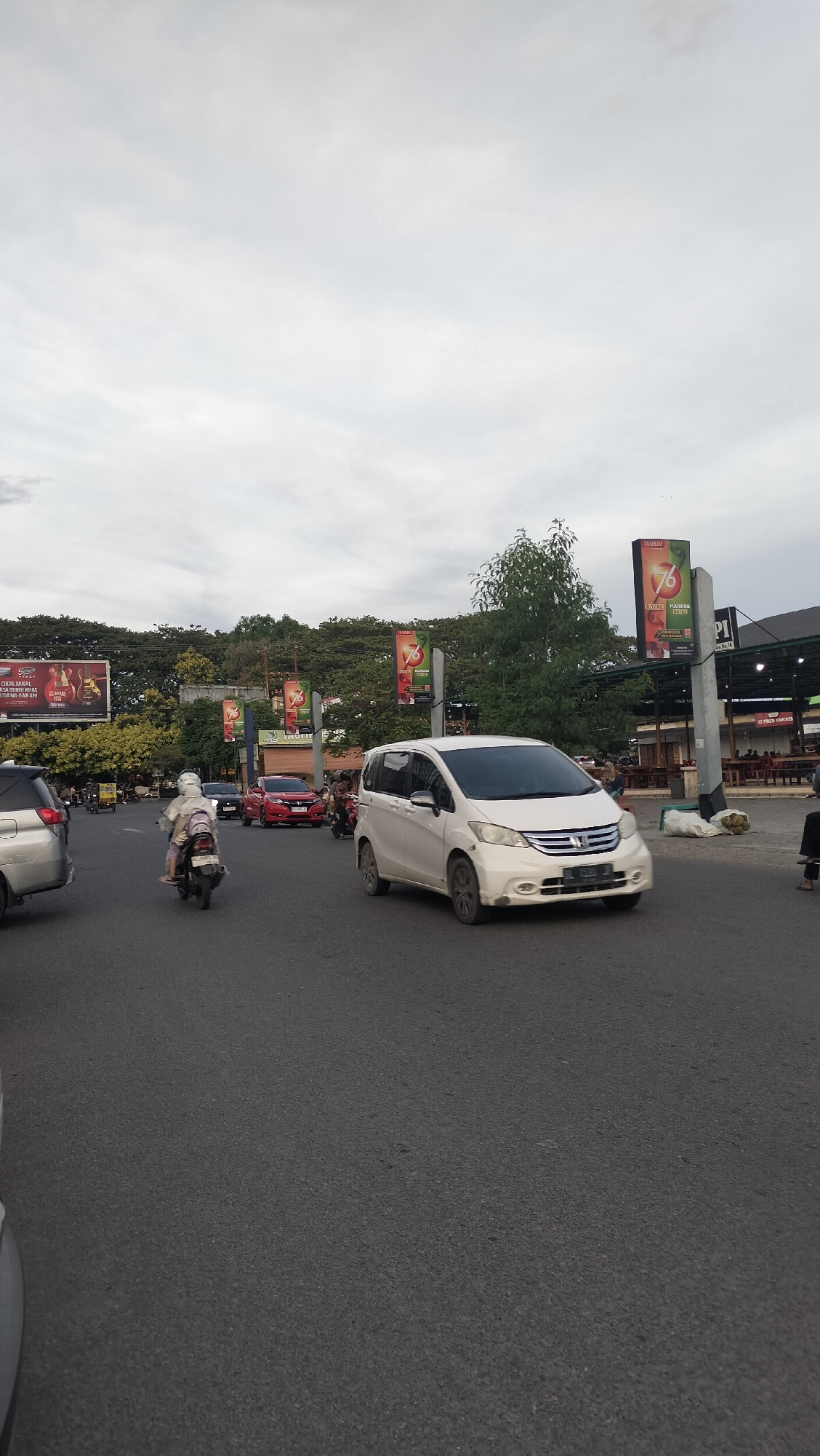 The image depicts a street scene in Banda Aceh, Indonesia, under an overcast sky. A white minivan, possibly a Honda Freed, is the most prominent vehicle in the foreground, driving on the right side of the road. To its left, a silver car is partially visible, also on the right side of the road. A person on a motorcycle, wearing a white helmet and a light-colored outfit, is moving away from the viewer on the left side of the road. Further down the road, a red car is seen. On the right side of the road, there is a building with outdoor seating and tables, suggesting a food establishment. Several tall, slender poles with red and white banners are erected along the roadside, some featuring the number "76". Trees and vegetation form a backdrop behind the banners and buildings. Billboards are visible in the distance on the left side of the image. The road surface is asphalt and appears to be in good condition. The overall atmosphere suggests moderate traffic during daytime.