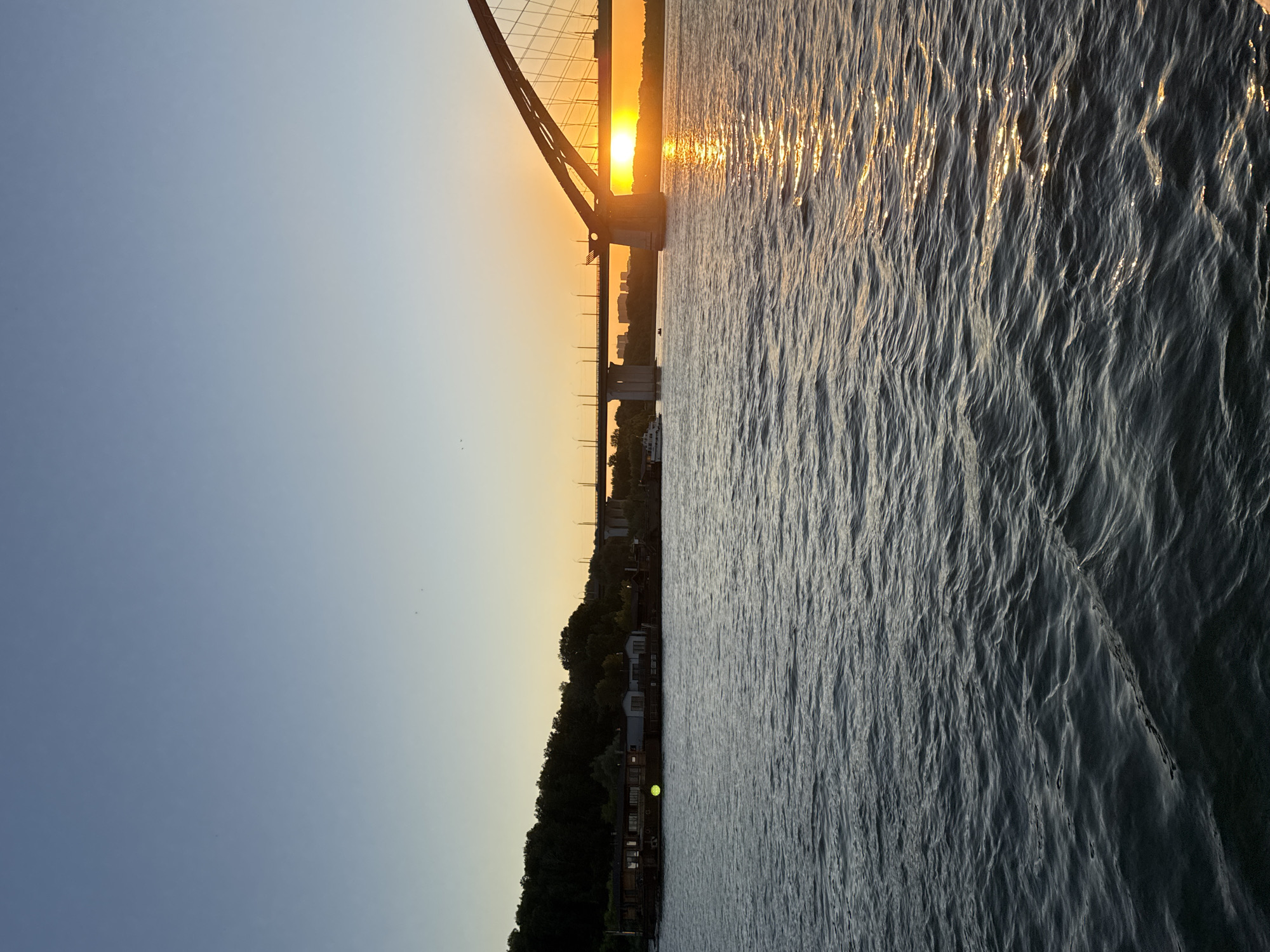 The image shows a sunset over the Ob River in Novosibirsk, Russia. The sun is setting behind a large suspension bridge, casting a warm glow across the sky and reflecting on the rippling water. The bridge, with its intricate metalwork and concrete supports, dominates the left side of the frame. Silhouetted against the sunset are the distant city skyline and a treelined shore. A boat is visible on the river, and a few buildings are nestled amongst the trees on the bank. The water is choppy, with sunlight glinting off the waves. The overall mood is peaceful and serene, capturing the beauty of a summer evening. The time of day is clearly sunset. The weather appears to be clear, with a gentle breeze indicated by the waves on the water.
