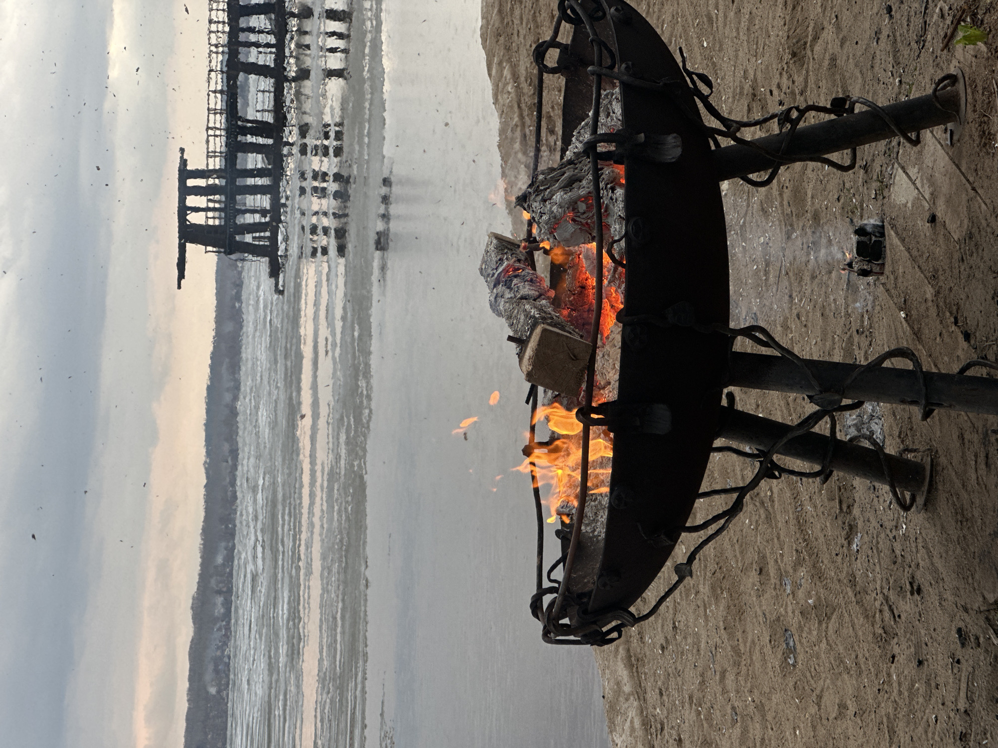 A black metal fire pit, containing burning wood, sits on a sandy beach next to a body of water. In the background, a dilapidated pier or structure extends into the water. The sky is overcast. The location is in Votkinsk, Russia.