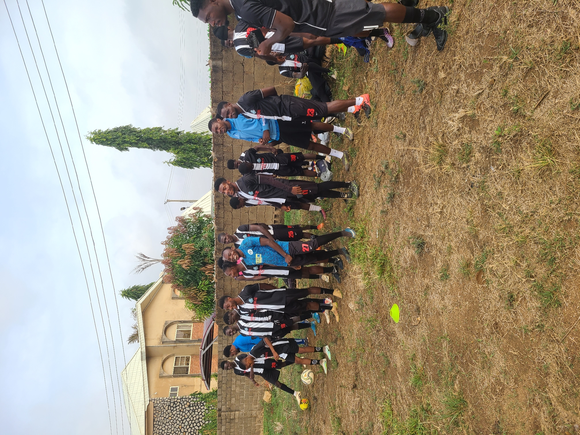A soccer team stands on a dirt field, lined up side-by-side in a line that stretches from the foreground to the background of the shot. Most of the team are wearing black and white striped jerseys and black shorts, and some wear blue jerseys. A soccer ball and a few yellow cones are on the ground. A brick wall is visible in the background behind the team. Behind the wall is a building that is surrounded by a tall, green tree. Overhead, the sky is overcast.
There is not enough information available in this image to confirm the location in the city of Bukuru, Nigeria.