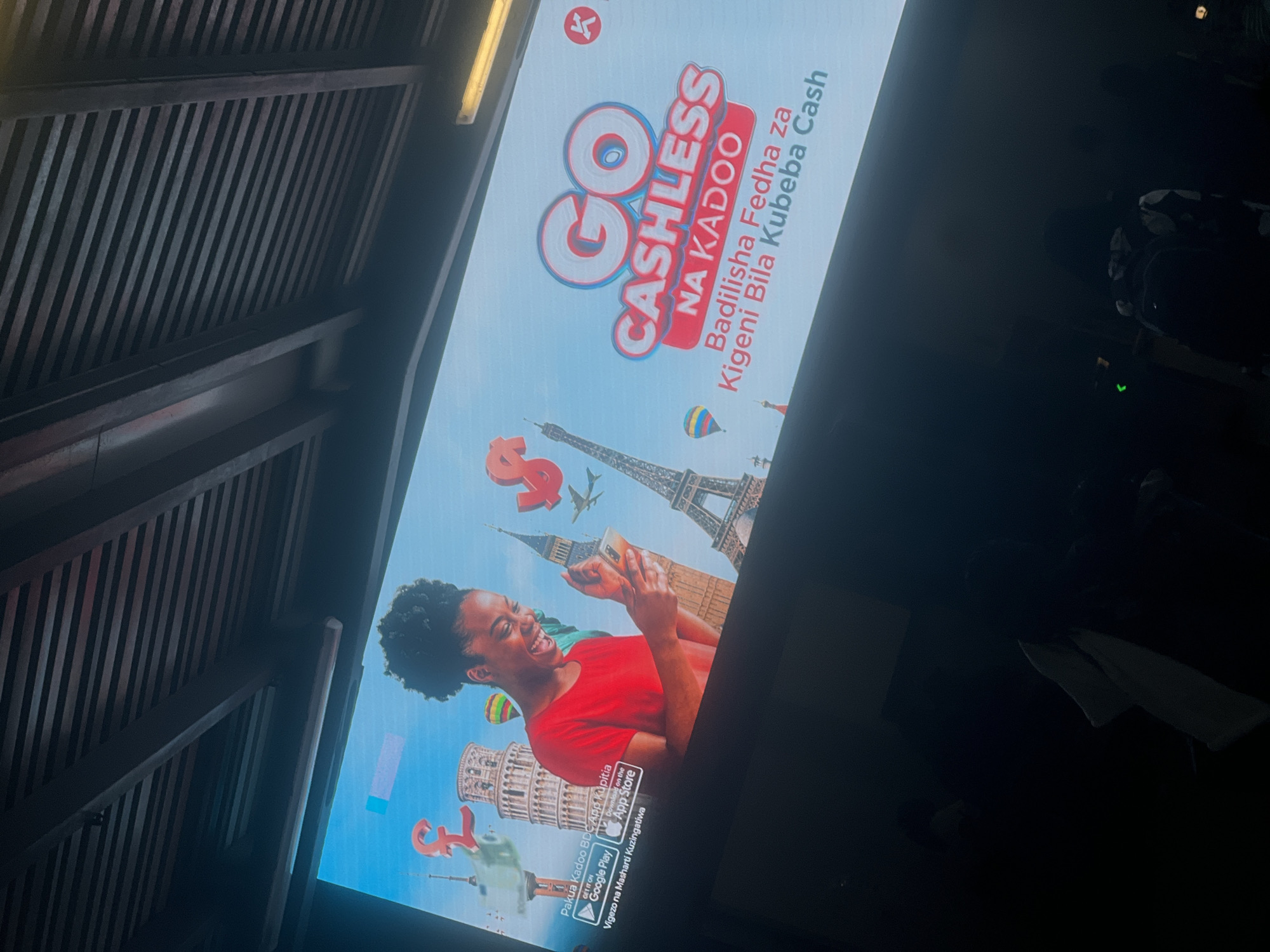 Here is a description of the image:
The image shows an advertisement for the "Go Cashless Na Kadoo" app, a digital finance platform in Tanzania. The advertisement is displayed on a screen with a dark wooden ceiling overhead.
The advertisement features a woman in a red shirt holding a smartphone. The background of the advertisement includes images of famous landmarks like the Eiffel Tower, the Leaning Tower of Pisa, and Big Ben, as well as a large dollar sign. The text on the ad is in Swahili and English, promoting the app as a way to exchange foreign currency without carrying cash. The logos for the Google Play Store and Apple App Store are also displayed. The "K" logo refers to Kadoo.
The overall scene suggests the advertisement is in an indoor location, likely a waiting area, lounge, or public space within a building or structure with a wooden ceiling. The presence of other people is visible in the background to the right, although their appearance is obscured by shadows.