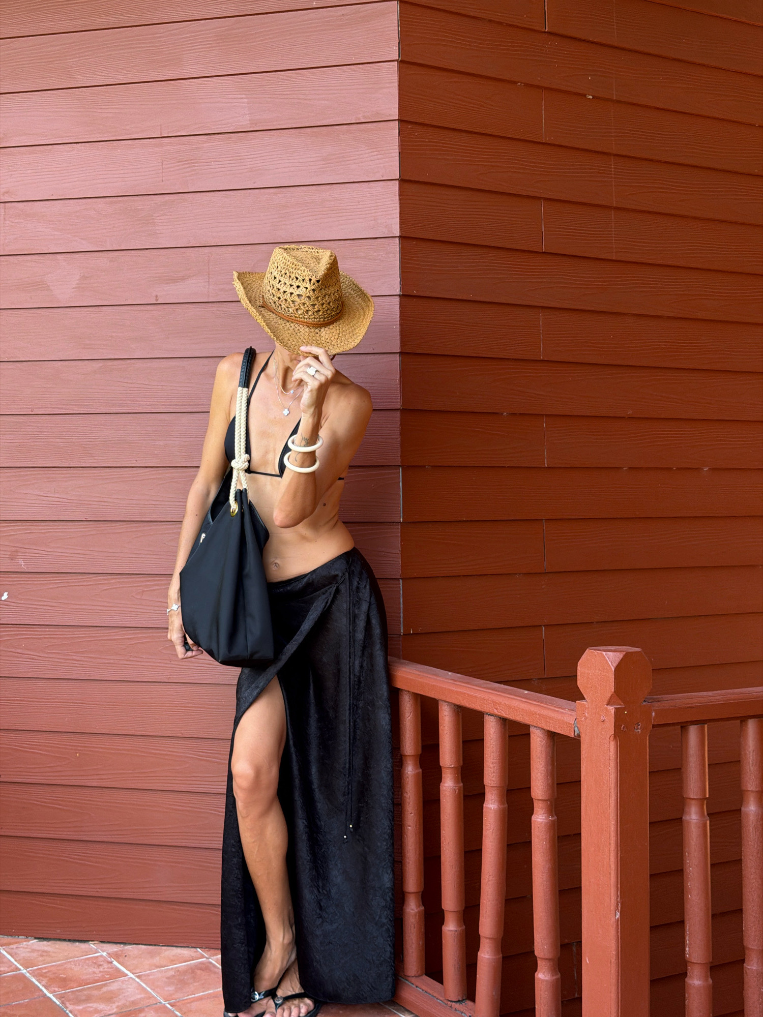 A woman wearing a straw hat, black bikini top, and a long black skirt with a high slit stands leaning against a reddish-brown wooden wall in Ko Samui District, Thailand. She holds a black tote bag on her left shoulder and her right hand is near her face, partially obscuring it. She wears several bracelets and necklaces. To the right of the woman is a red wooden railing with vertical posts. The ground beneath her feet is tiled.