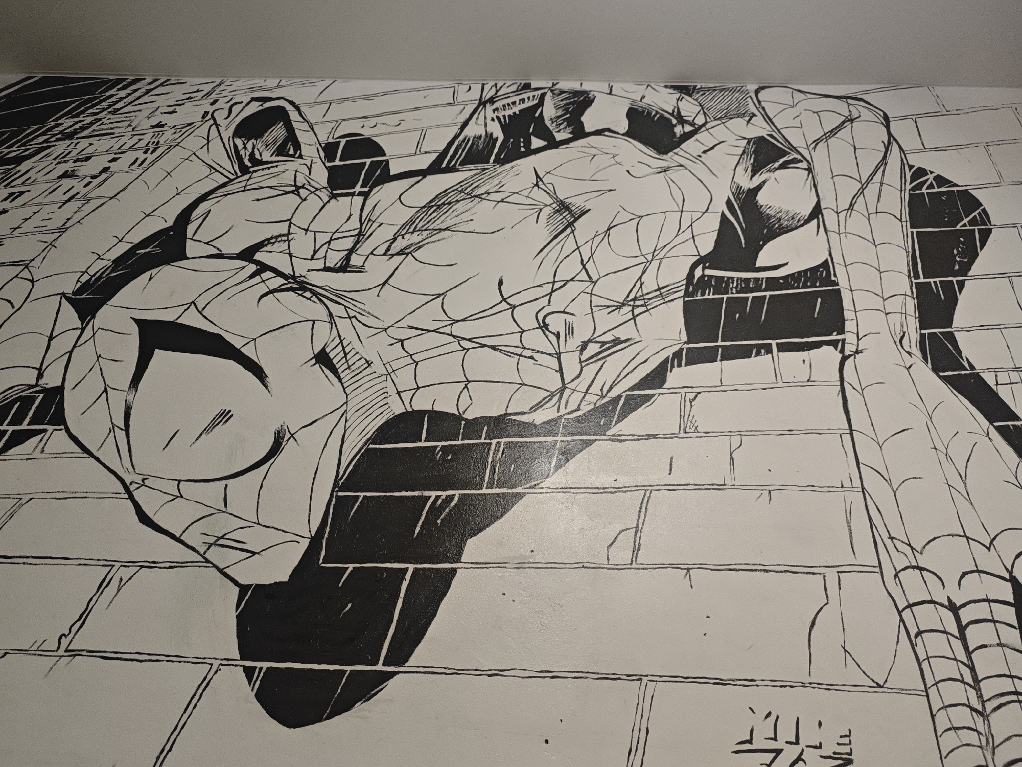 A black and white mural depicting Spider-Man is painted on a brick wall. The superhero is shown in a dynamic pose, seemingly hanging or crawling, with his web pattern clearly visible on his suit. The artwork utilizes bold lines and shading to create a sense of depth and movement. The brick pattern of the wall is also depicted, with grout lines and some imperfections adding to the texture. The mural is located in the city of O'lkhovka, Russia.
