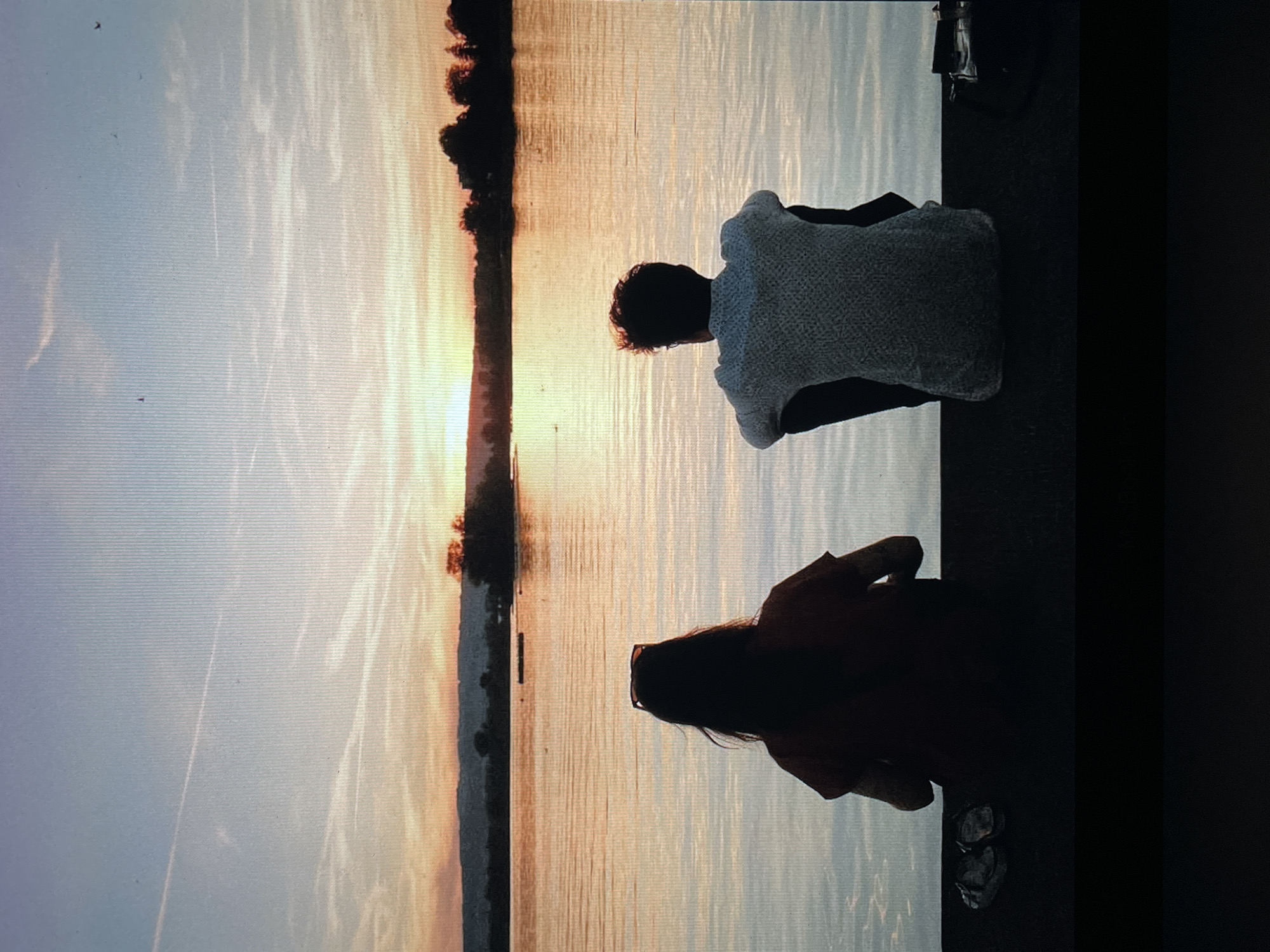 Two people sit near a body of water at sunset. The scene features a shoreline with trees silhouetted against the fading light. The exact location in Saint Petersburg, Russia is undetermined from the photograph alone, but the scene could depict the Neva River or one of the city's canals. One person is wearing a light-colored sweater, and the other is wearing darker clothing. Their activities are observational, seemingly watching the sunset over the water. Sandals are visible near one individual.