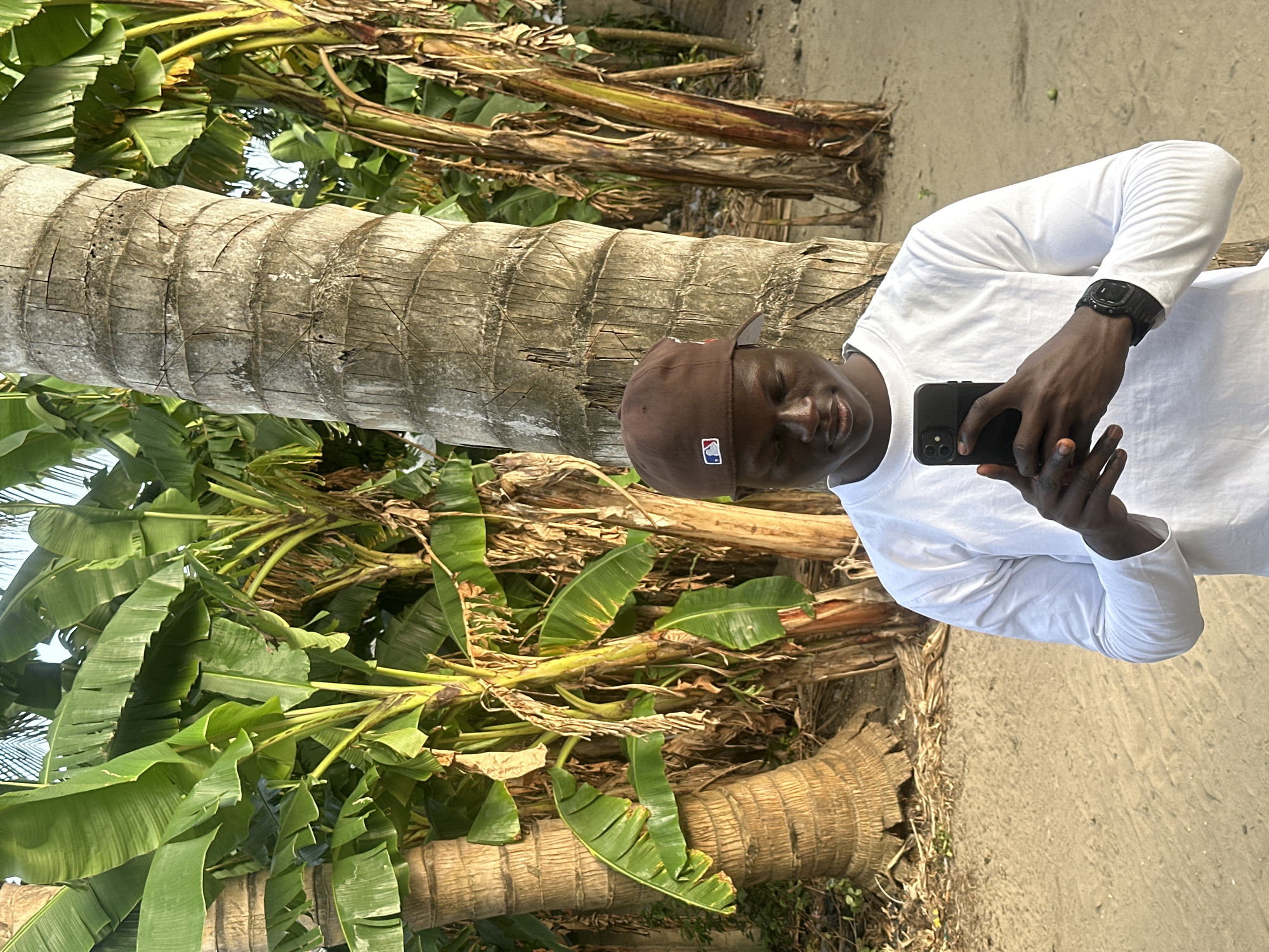 In the city of Badagry, Nigeria, a young Black man wearing a white long-sleeved shirt and a brown baseball cap holds a black smartphone, taking a photo of himself. He stands between two large palm trees with a background of lush green banana trees. The ground is sandy and dry, with some scattered leaves and debris. The man's face is partially obscured by the phone as he looks down at it. A black digital watch is visible on his left wrist. The lighting suggests it is either early morning or late afternoon.