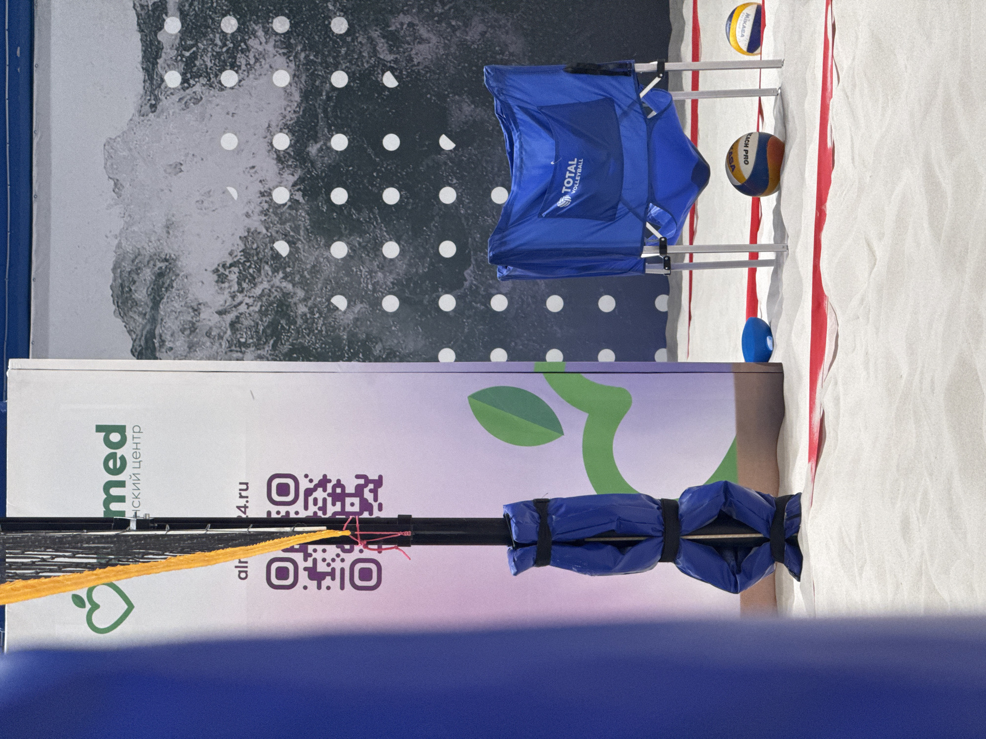 The image shows a beach volleyball court. A blue beach chair with "TOTAL VOLLEYBALL" printed on it sits near the edge of the court, along with two volleyballs (one Mikasa) and a blue marker. A banner behind the net has "med" and "медицинский центр" printed on it, along with a QR code and a logo featuring a green leaf. The banner also has "alr24.ru". The court is sandy with white markings. The location is likely Krasnoyarsk, Russia, based on the Cyrillic text on the banner.