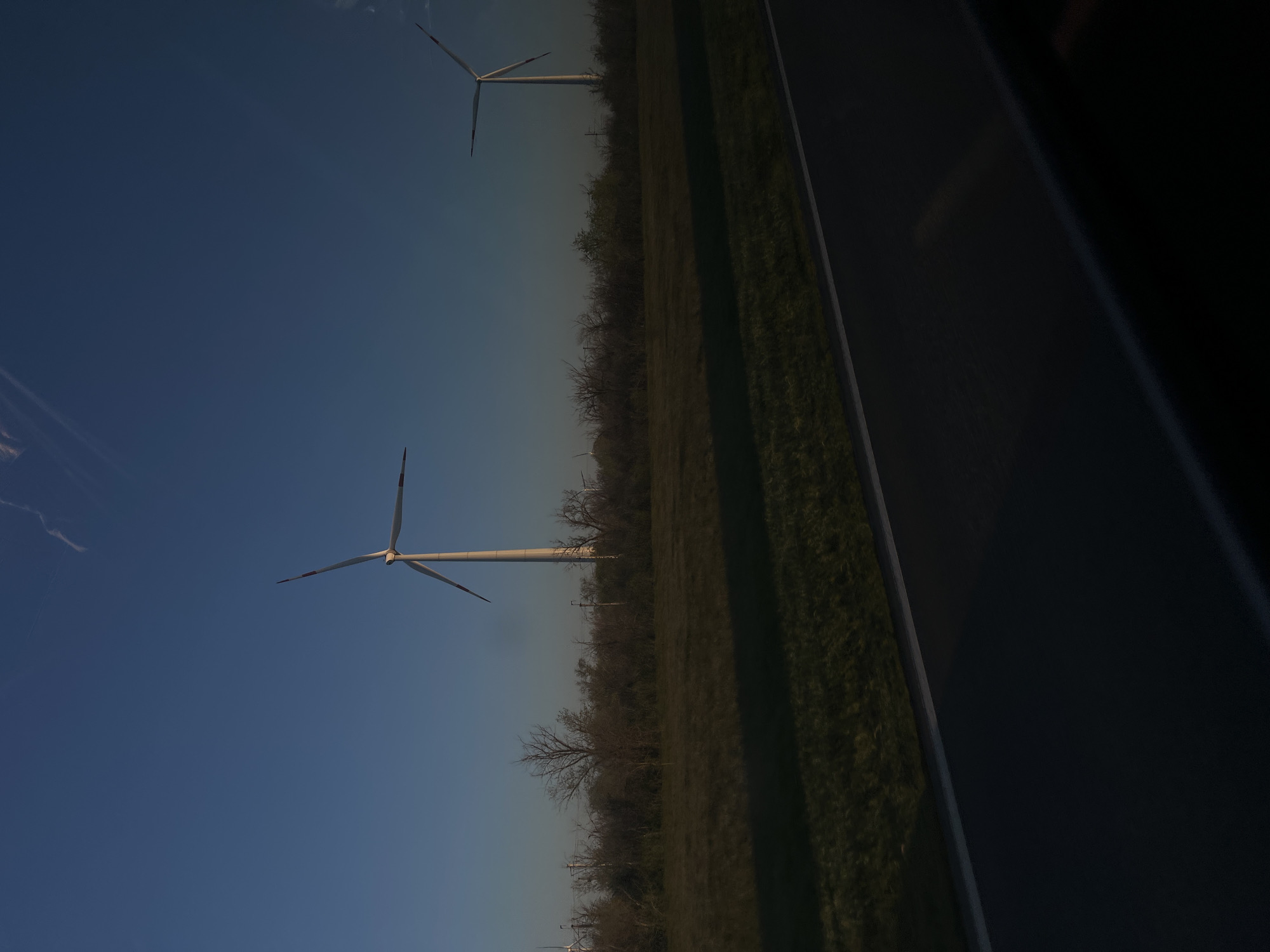 The image depicts a view from inside a moving vehicle, likely a car, with the landscape seen through the window. Two wind turbines are visible against a clear blue sky. One turbine is closer and appears larger in the frame, while the other is smaller and further away. A field or grassy area and a line of trees or shrubs run parallel to the road or surface the vehicle is traveling on. A portion of the vehicle interior, possibly the dashboard or window frame, is visible on the right side of the image. The location is assumed to be in Zimovniki, Russia, as requested.