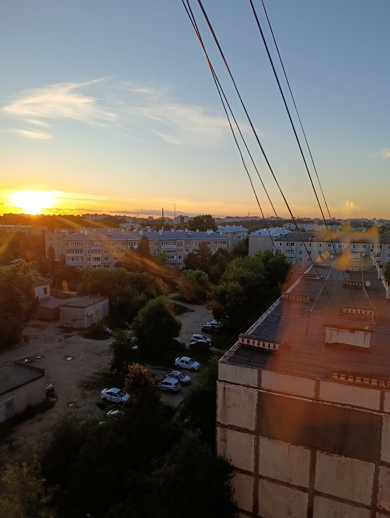 This is an aerial, eye-level, medium shot of a city skyline at sunset in Alexandrov, Russia. The sky is a gradient of light blue to orange, with wispy clouds. The sun, a bright orb of light, is setting on the left side of the frame, casting long shadows and lens flares across the scene. In the foreground, several black electrical wires stretch diagonally across the upper portion of the image.
The middle ground is dominated by apartment buildings, several of which are tall, multi-story structures with visible windows and balconies. Some of these buildings appear to be made of concrete and have a gray or pale blue facade. Interspersed between the buildings are dense clusters of green trees.
In the lower left quadrant of the image, there is a parking lot with several cars visible. The cars are various colors, including white and dark gray. There are also some smaller, low-lying buildings or sheds in this area, some with brick or concrete exteriors.
The bottom right quadrant of the image features the rooftop of a large, multi-story building, likely an apartment block. This rooftop is flat and dark, with visible conduits and what appear to be satellite dishes. The side of this building, visible at the very bottom of the frame, is made of concrete blocks and exhibits some signs of wear. The overall impression is of a residential urban environment bathed in the warm light of the setting sun.