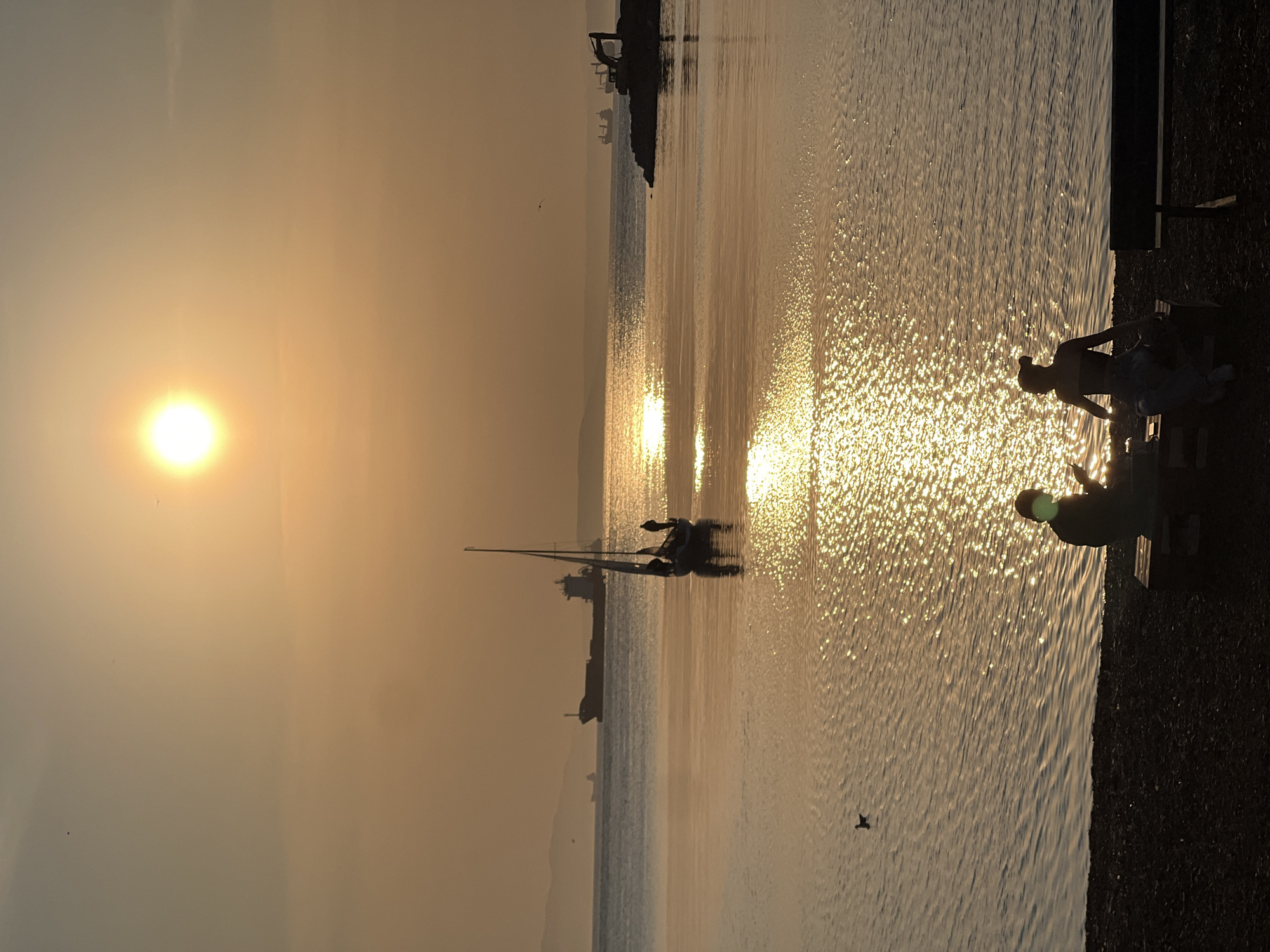 The image shows a sunset over the water in Vladivostok, Russia. The sun is low in the sky, casting a golden glow over the scene. The water is calm, and reflects the sunlight. There are boats in the distance, and people are sitting on a bench on the right.