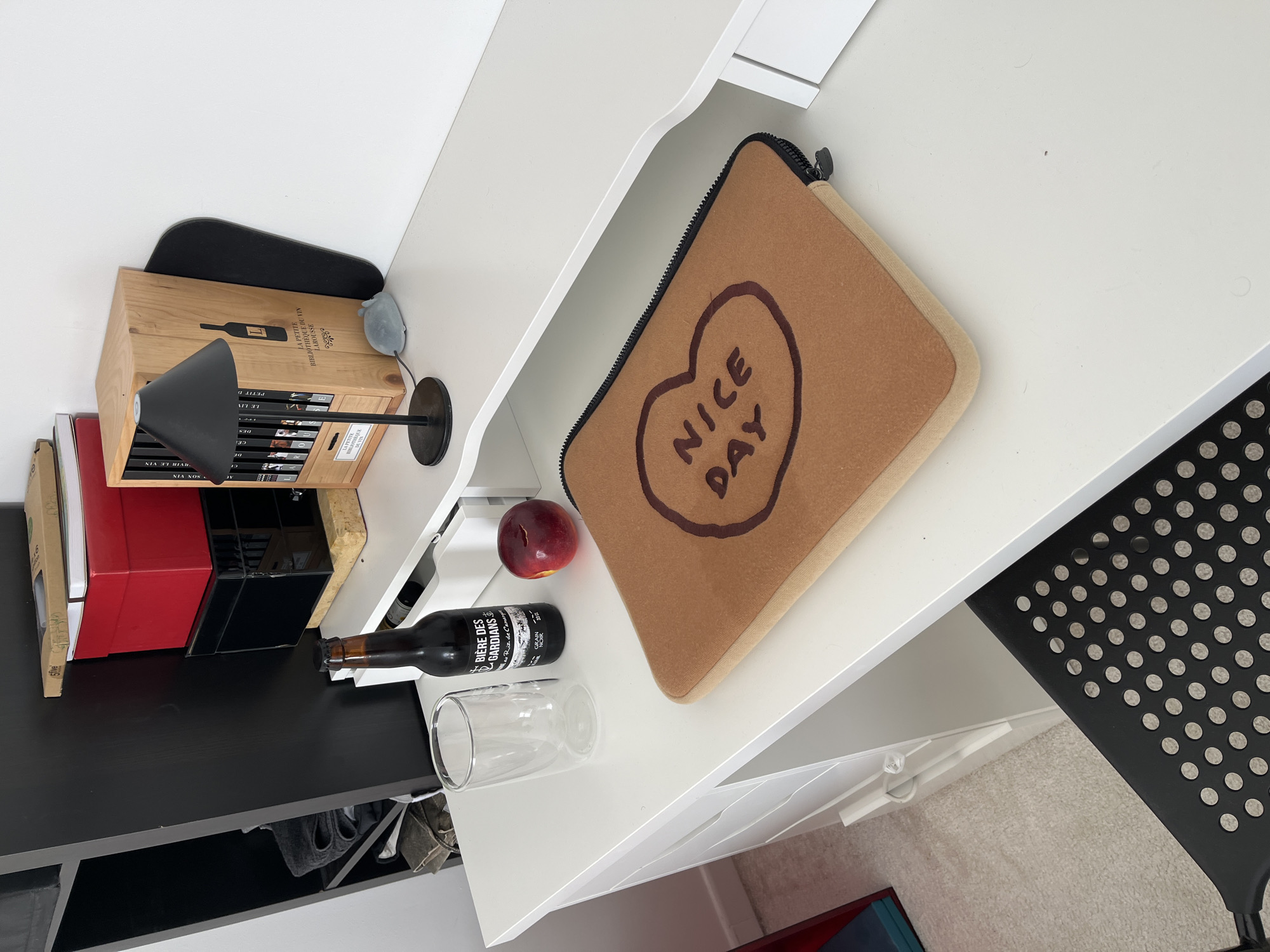 The image depicts a desk with various items. A brown laptop case with a heart graphic and the words "NICE DAY" lies on the desk. Next to it is a red plum and a glass bottle of "BIÈRE DES GARDIANS" next to a glass. A wooden box labeled "LA PETITE BIBLIOTHÈQUE DU VIN LAROUSSE" is also present, along with a small black lamp and books. A black chair with circular cutouts is partially visible in the lower right corner. The scene is set indoors, with a combination of white and black furniture. There is no indication of the location being Chambœuf, France.