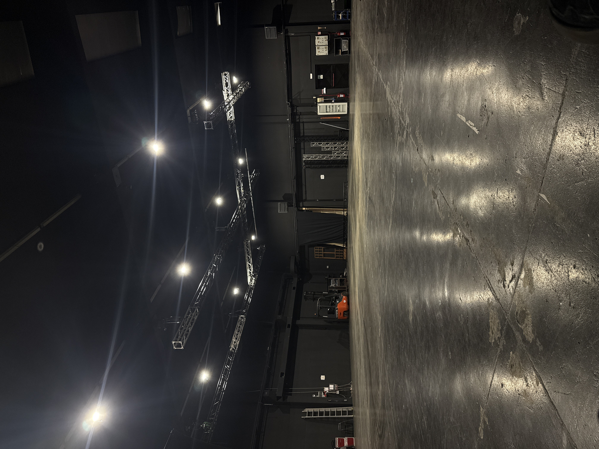 This image captures a large, unoccupied studio or production facility located in Cologno Monzese, Italy. The space features a dark gray concrete floor, showing visible scuffs, wear, and light-colored patched areas, with prominent reflections from overhead lighting. The walls and ceiling are painted a uniform matte black.
An extensive grid of silver metal trusses is suspended from the ceiling, supporting numerous bright white stage or studio lights that illuminate the room. Several ventilation grilles are affixed to the upper sections of the walls.
Along the left wall, a silver step ladder leans against the surface, flanked by two red equipment cases and a black chair. Electrical conduits and a mounted control panel are also present in this area.
Centered against the back wall, an orange Toyota forklift is parked, facing left. To its right, a large section of black theatrical drapery is installed, adjacent to vertically stacked wooden stage flats or frames.
The right side of the back wall includes a stack of metal trusses, a white upright cabinet or appliance, two red fire extinguishers mounted near dark doors (one marked with a red 'EXIT' sign), and multiple electrical control panels. A blue rolling utility cart is partially visible on the far right. No individuals, actions, or interactions are depicted in the static scene.