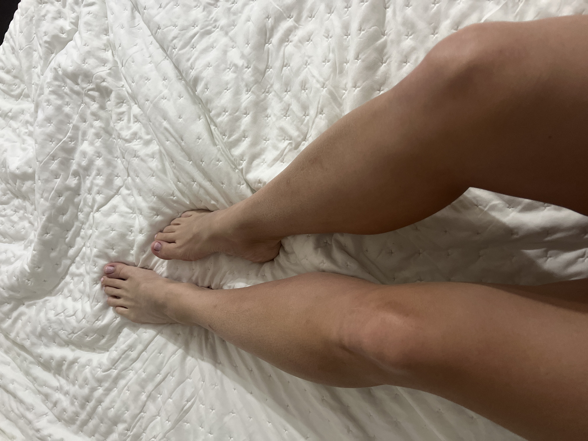 The image displays a first-person, downward view of an individual's bare lower legs and feet resting upon a white, quilted bedspread. The bedspread features a uniform pattern of small, embossed cross-shaped stitches. The toenails on both feet are painted with a pale pink polish. The skin on the visible limbs exhibits a light tan complexion. No other individuals or objects are present within the frame. The scene is located indoors in Budva, Montenegro.