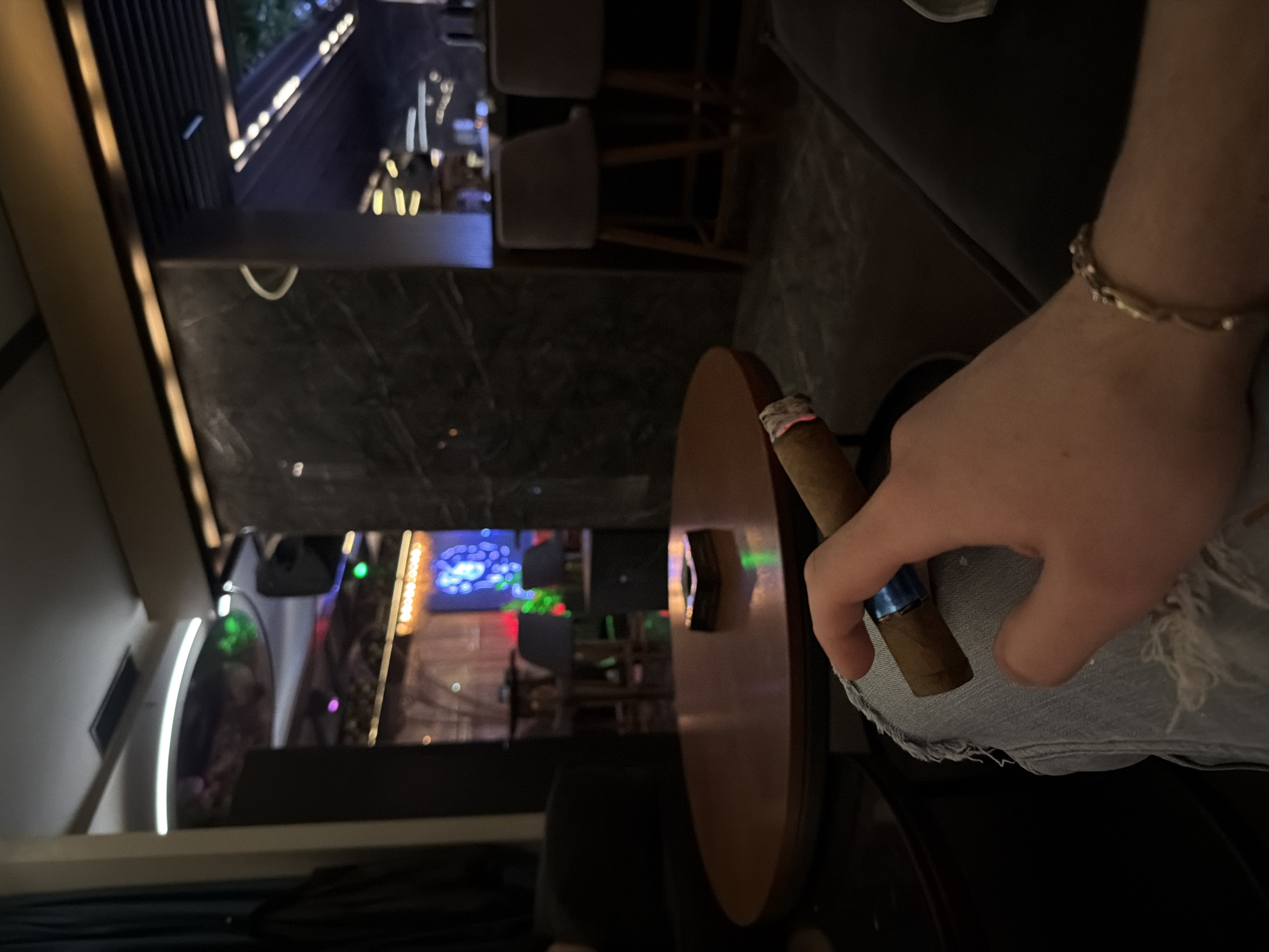 A rotated eye-level shot captures a scene in an indoor setting, possibly a bar or lounge, featuring a person's hand holding a cigar. The hand is positioned over a light-wash denim material with distressed edges, presumably part of the person's clothing. A simple gold bracelet adorns the wrist. The cigar has a dark band around it. A round wooden table is partially visible next to the hand. In the background, a dimly lit interior with furniture, a bar area, and decorative lighting is seen. A cityscape with bright lights, possibly from buildings or billboards, is visible through a window or opening in the background.