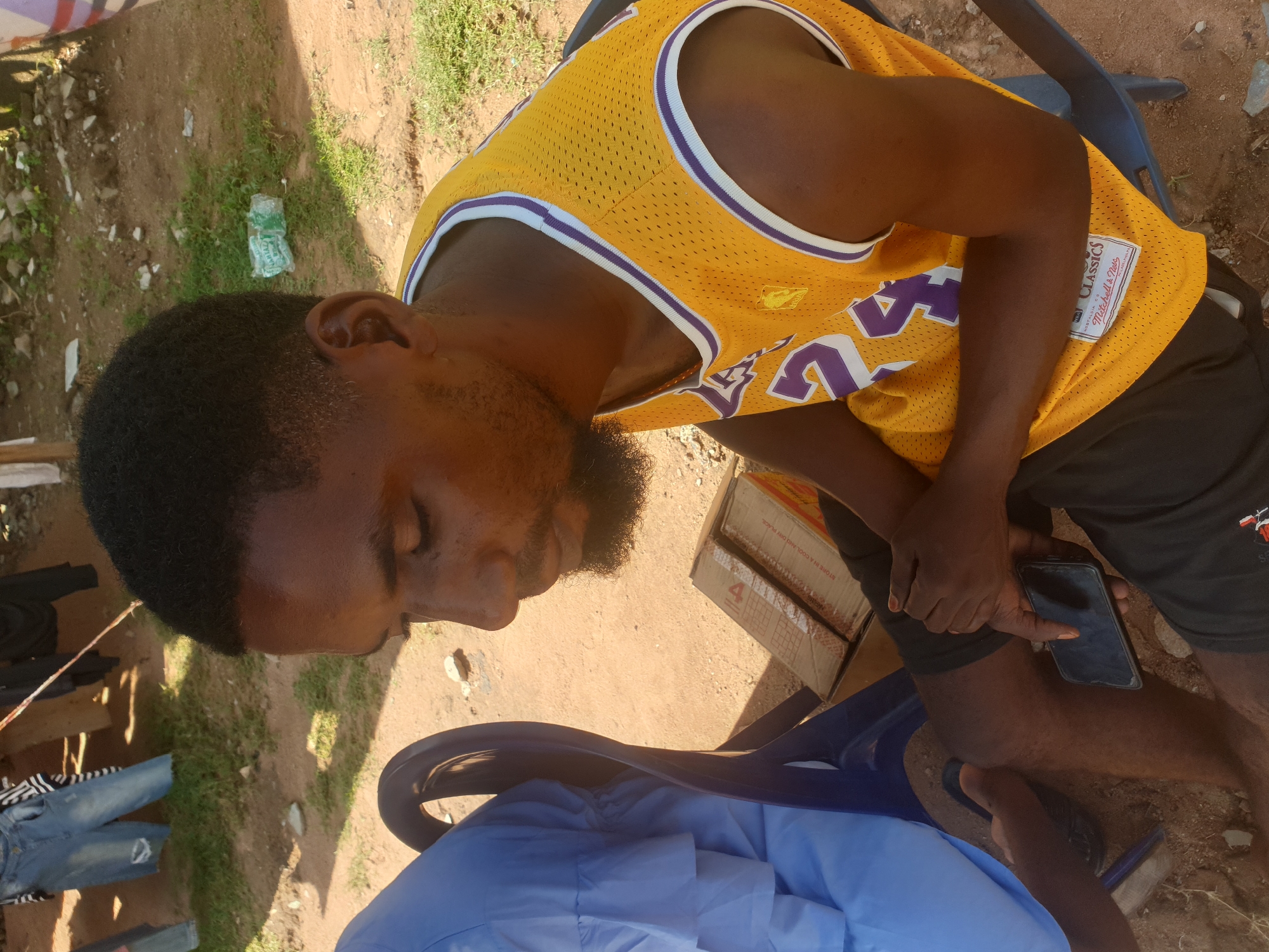 The image captures two individuals in an outdoor setting, likely in the city of Jalingo, Nigeria. The person in the foreground is a dark-skinned male, wearing a yellow and purple basketball jersey with the number 24 prominently displayed. He has short, dark hair and a beard. He is holding a dark-colored smartphone in his right hand and appears to be looking downwards. His left hand is resting on his knee. The person to his left, mostly obscured by the foreground individual and a blue plastic chair, is wearing a light blue garment with ruffled sleeves. The environment appears to be a dusty, outdoor area with patches of dry grass and scattered rocks. In the background, denim jeans are hanging on a line, suggesting a domestic or informal setting. A crumpled plastic bottle is visible on the ground to the right of the foreground subject. The overall lighting suggests it is daytime.