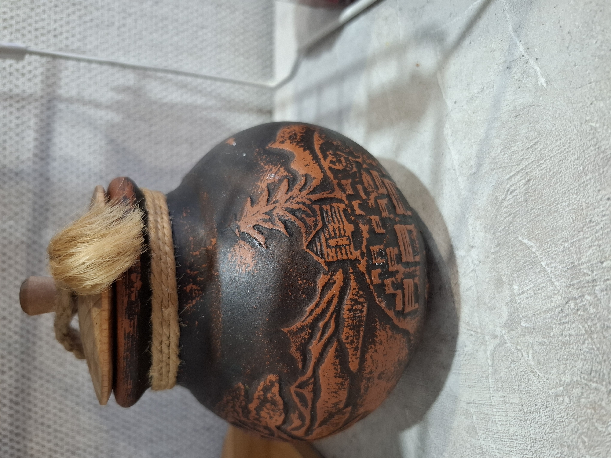 The image shows a spherical, clay-like vessel with a dark brown/black body and orange-brown decorations. The decorations appear to be embossed or etched into the surface, depicting patterns that may represent natural elements or architectural structures. The vessel has a wooden lid secured with twine and a bundle of straw-like material. The background features a textured, light-colored wall. The image does not provide specific information about the location, people, subjects, actions, or interactions. The image also doesn't provide information about the city of Амзя, Russia.
