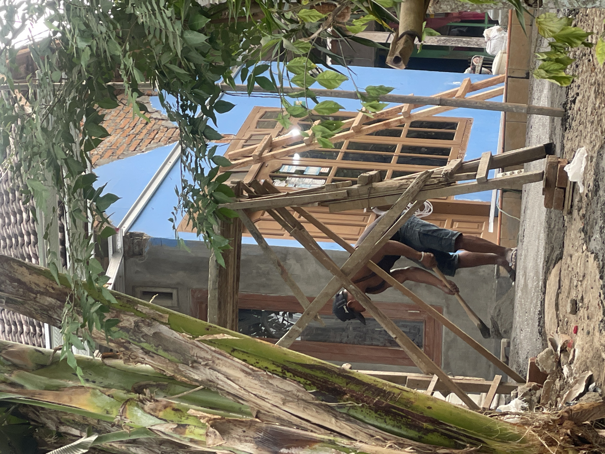 In Parakan, Indonesia, a man wearing a black baseball cap, a dark sleeveless shirt, and denim shorts is engaged in construction work. He is using a shovel to move a pile of grey material, likely cement or gravel, on a dirt and concrete surface. The scene is outdoors, with a blue building under construction in the background, featuring wooden window frames. Scaffolding made of wooden planks is erected around the building. Lush green foliage from trees frames the construction site, partially obscuring the view. The ground is littered with bricks, debris, and construction materials.
