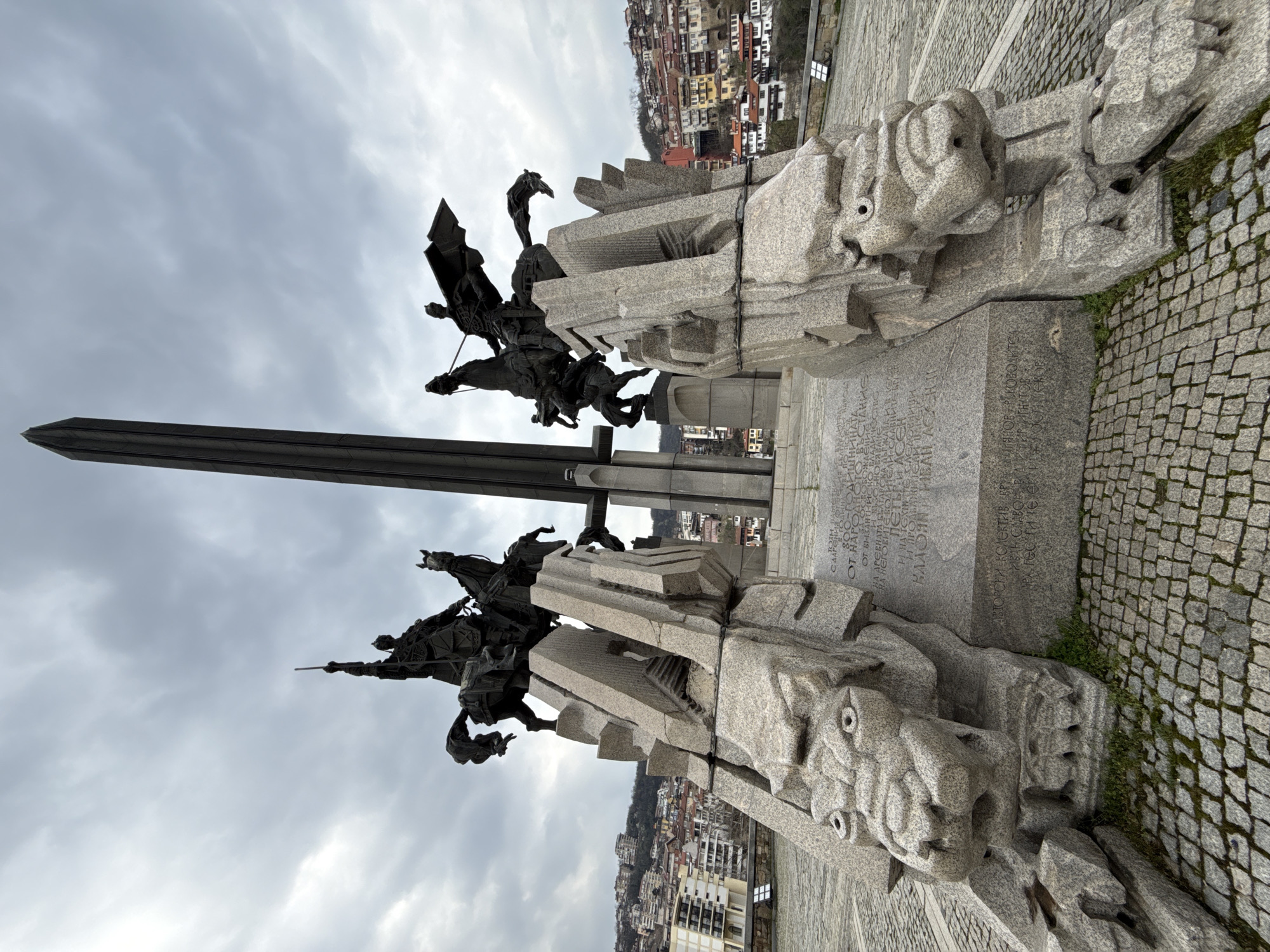 This image depicts the Monument to the Asen Dynasty, a large, abstract monument featuring bronze equestrian statues on top of granite blocks with lion head carvings, located in Veliko Tarnovo, Bulgaria. The sky is overcast and the city is visible in the background. The inscription at the base of the monument is in Cyrillic script.