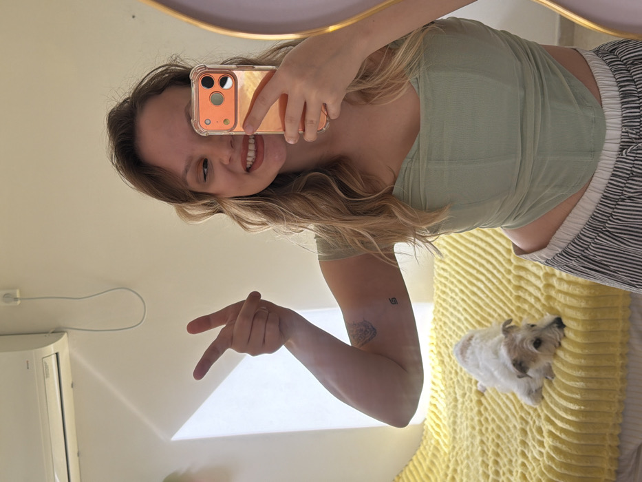 An indoor mirror selfie depicts a young woman in Tel-Aviv, Israel. The woman, with wavy light brown hair, wears a light green corset-style top and striped pants. She smiles, partially revealing teeth, and holds an orange-backed smartphone with her right hand, obscuring the right side of her face. Her left arm is raised in a peace sign, displaying two tattoos on her bicep and forearm. To her left, on a bed covered by a bright yellow textured blanket, a small white and brown terrier-type dog rests, facing left. The background features a light-colored wall with a white split-unit air conditioner mounted in the upper left, connected by a visible power cord. Natural light enters from the left, casting a diagonal shadow.