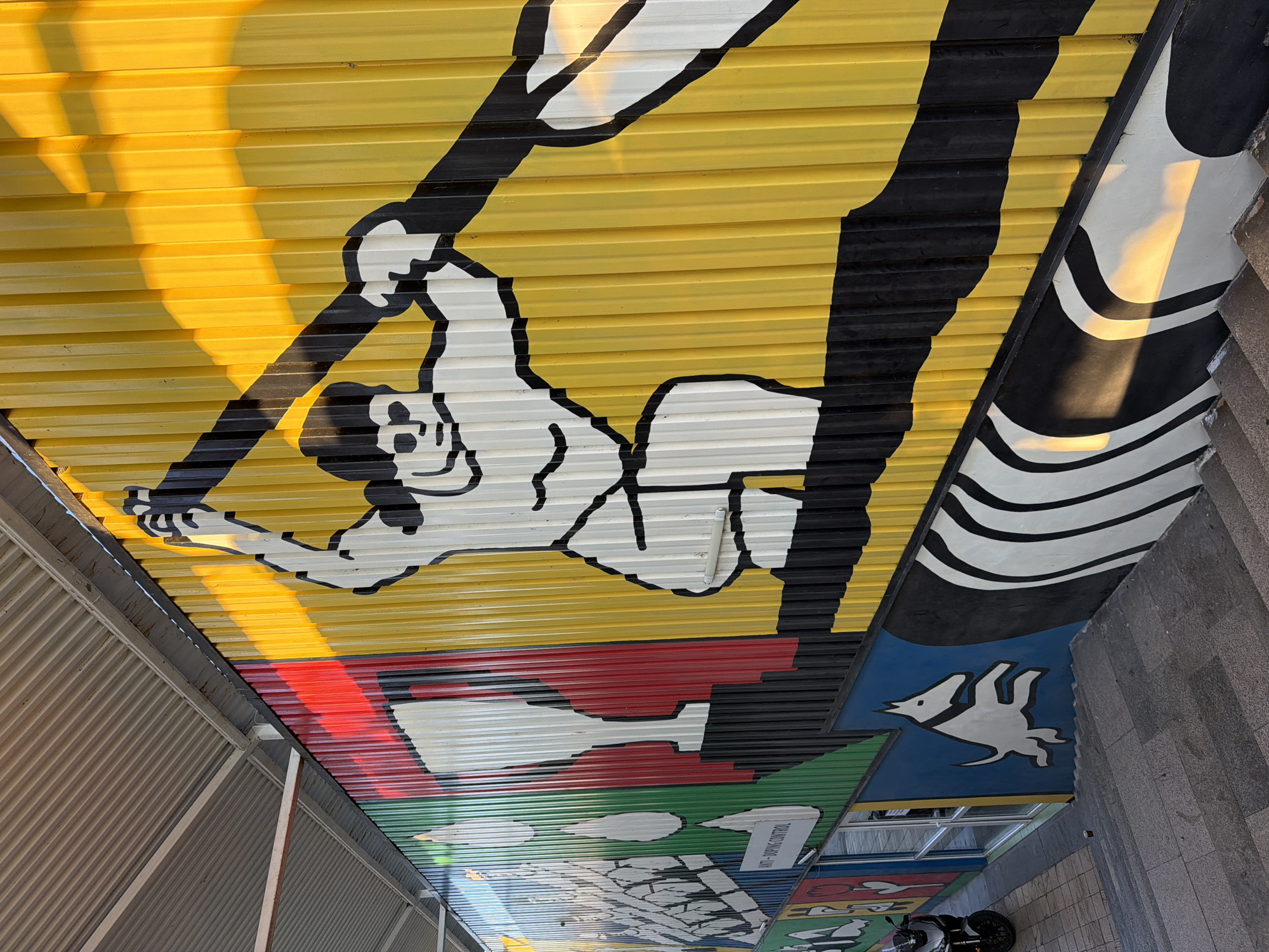 The image shows a brightly colored mural on a corrugated metal wall in Plovdiv, Bulgaria. The mural depicts a stylized figure in the center, appearing to be running or in motion, with a large, dark diagonal line suggesting a weapon or pole. To the left of this figure, there are abstract shapes and elements in red, green, and white, including what looks like a trophy and stylized drops. Further left, there are depictions of candles and possibly flowers. Below the central figure and on the left, a white dog with a blue background is shown in a leaping pose. The lower section of the wall features a black and white abstract design with wavy lines. The wall is illuminated by the sun, casting strong shadows and creating a warm glow in some areas. In the foreground, concrete stairs are visible, leading up towards the mural. A parked motorcycle is partially visible on the left side of the frame, near a glass-fronted doorway. The overall scene suggests an outdoor public space, likely a walkway or passage decorated with contemporary art.
