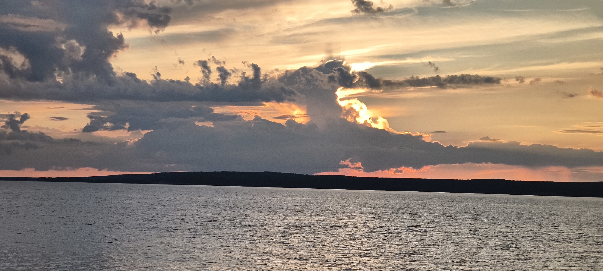 The image shows a wide shot of a body of water at sunset. The sky is filled with dramatic clouds, with the sun's rays breaking through, casting a warm orange and yellow glow. The clouds range in color from dark grey to lighter shades of pink and orange. A dark, silhouetted landmass, likely a forest or hills, forms a horizon line across the lower third of the image. The water is calm, with a slight ripple texture reflecting the colors of the sky. The location is described as Unknown, Unknown.