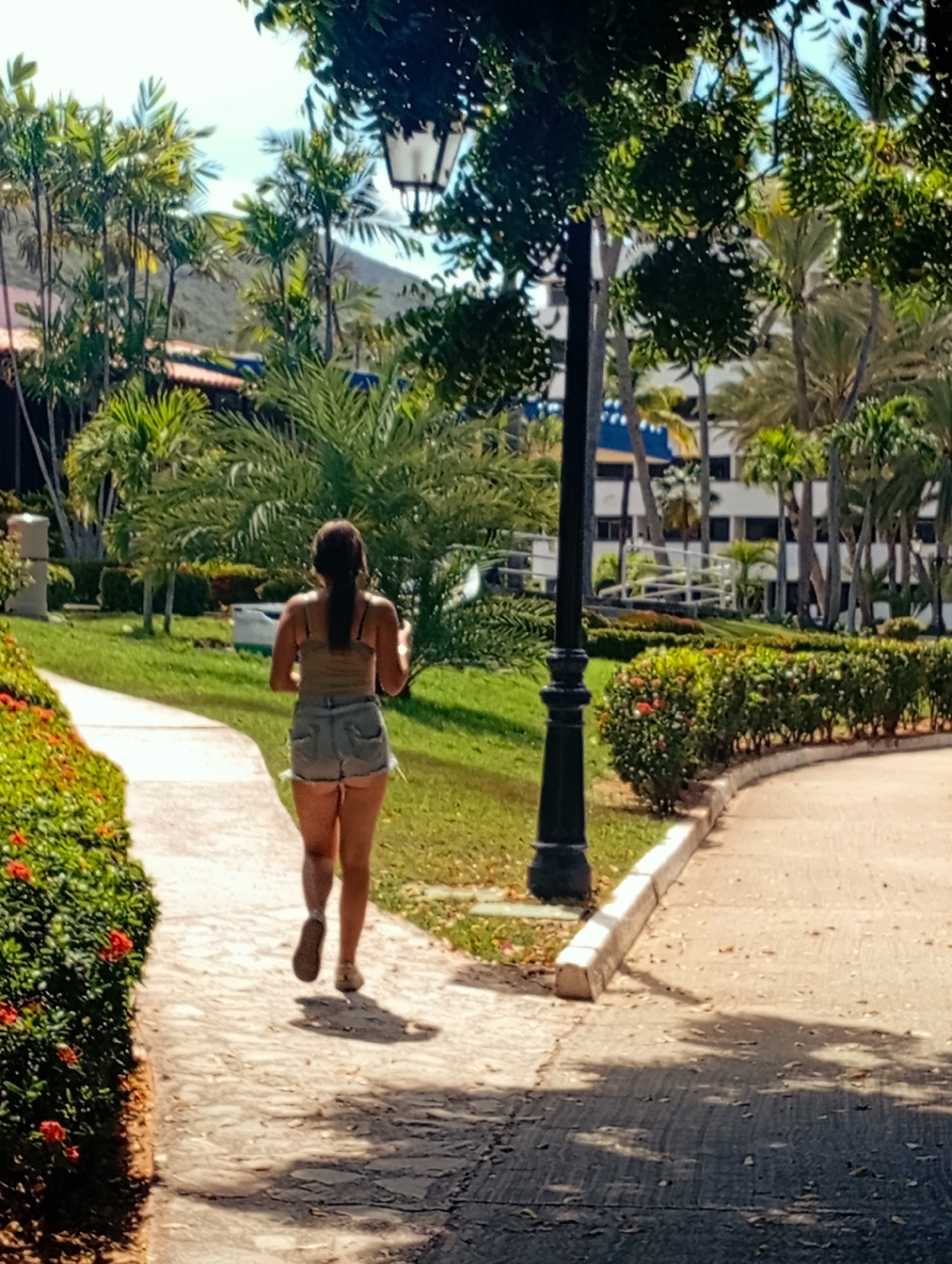 A woman walks away from the viewer on a paved path in a park-like setting in Playa El Agua, Venezuela. She is wearing denim shorts and a tank top. The path is lined with plants and flowers on one side, and a manicured hedge and a black lamppost on the other. In the background, there are palm trees, other trees, and a multi-story building. A mountain is visible in the far distance. The image appears to be taken during the day, with natural light and shadows on the path.