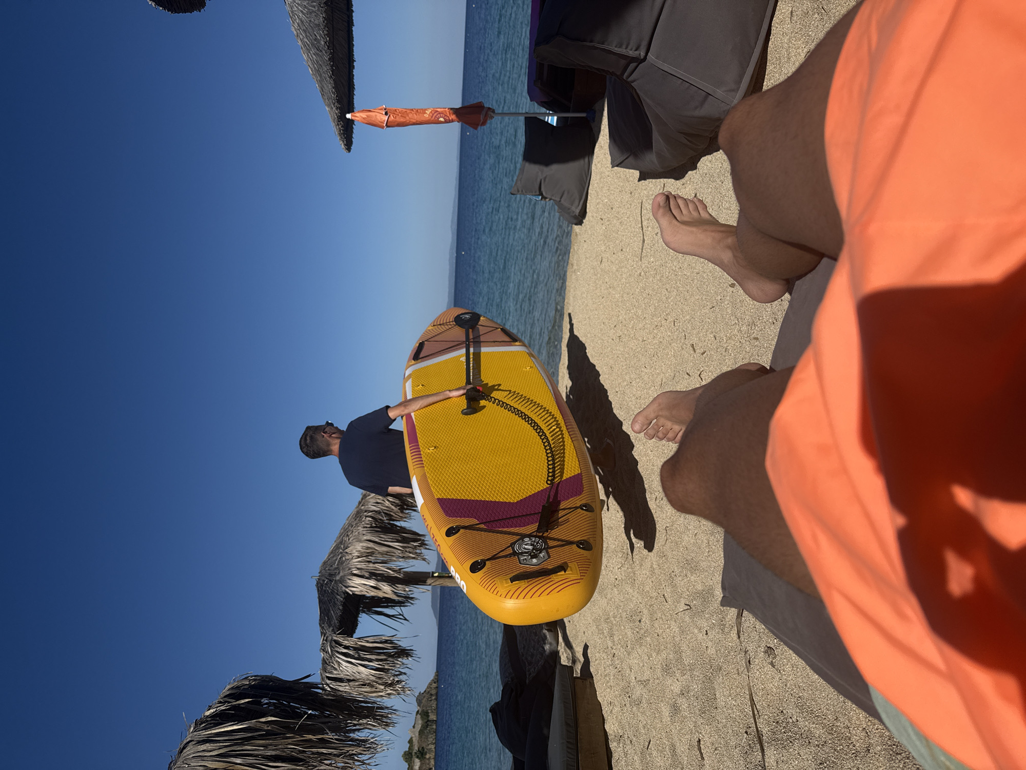 In Toroni Municipal Unit, Greece, on a sandy beach under a clear blue sky, a man carries a yellow and orange paddleboard towards the calm blue sea. He wears sunglasses and a dark t-shirt. The foreground shows the legs of a person relaxing on a beach lounger, wearing orange swim trunks. Beach umbrellas made of dried palm fronds and a modern grey beanbag chair are visible, suggesting a resort or leisure area. The water is calm, with a distant shoreline visible under the bright sun.