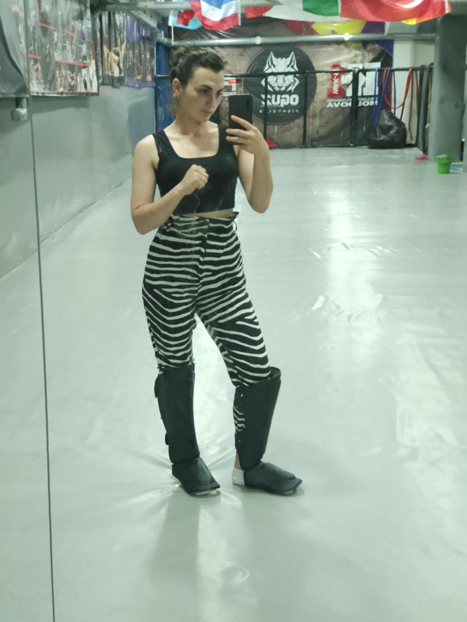 The image shows a woman in athletic gear posing for a mirror selfie in a gym. She is wearing a black crop top, zebra-striped pants, and shin guards with black foot coverings. The gym appears to be a martial arts or combat sports facility, as indicated by the banners with logos and flags hanging on the wall in the background. One banner displays a wolf's head logo with Cyrillic text. The floor is a light-colored, reflective surface, possibly a mat. The overall lighting suggests an indoor environment with overhead lights. The city of Comrat, Moldova, is mentioned as the location, but there are no specific visual cues within the image that definitively confirm this location.